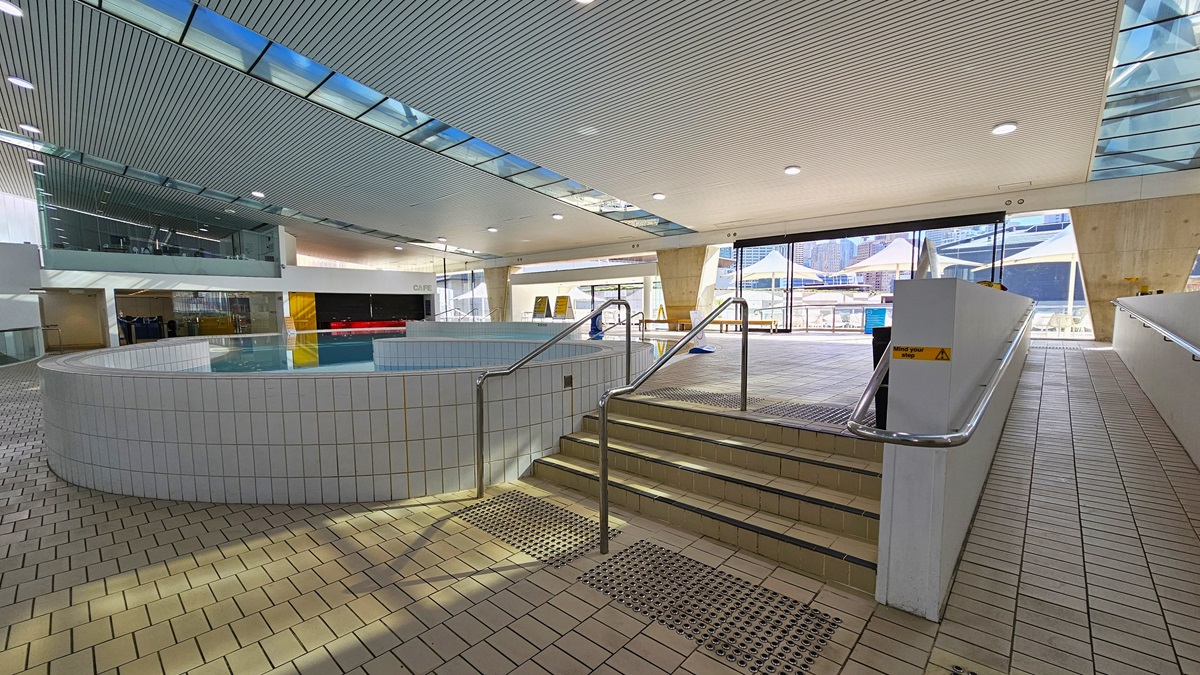 The Sauna Pool features steps with handrails and a ramp with handrails, along with truncated domes on the floor. Adjacent to it is the Program Pool, also equipped with steps and ramp entry, with visible safety signage. To the left, a glass wall provides a view of other facilities. The ground floor is tiled with small bricks, and the ceiling displays a white porous design with circular LED lights, incorporating clear glass reflecting the pool below. Beyond lie the sauna and Program Pool. The Sauna Pool features steps with handrails and a ramp with handrails, along with truncated domes on the floor. Adjacent to it is the Program Pool, also equipped with steps and ramp entry, with visible safety signage. To the left, a glass wall provides a view of other facilities. The ground floor is tiled with small bricks, and the ceiling displays a white porous design with circular LED lights, incorporating clear glass reflecting the pool below. Beyond lie the sauna and Program Pool.