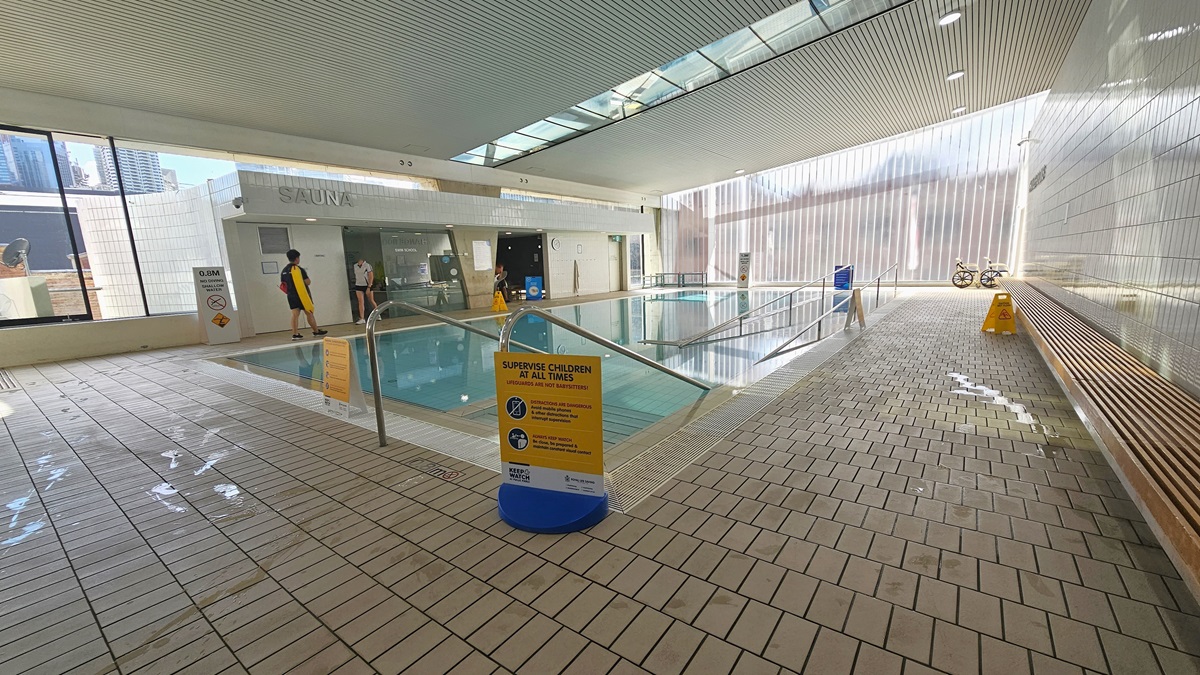 The Program Pool is furnished with steps and a ramp with handrails, accompanied by a ramp with handrails. Freestanding banners are affixed to the pool deck. A long wooden bench is situated against the right wall, and aquatic wheelchairs are positioned in the corner. The ground floor is tiled with small bricks, and the ceiling exhibits a white porous design with circular LED lights, featuring clear glass glass reflecting the pool below. Beyond lies the sauna and Program Pool. The Program Pool is furnished with steps and a ramp with handrails, accompanied by a ramp with handrails. Freestanding banners are affixed to the pool deck. A long wooden bench is situated against the right wall, and aquatic wheelchairs are positioned in the corner. The ground floor is tiled with small bricks, and the ceiling exhibits a white porous design with circular LED lights, featuring clear glass glass reflecting the pool below. Beyond lies the sauna and Program Pool.