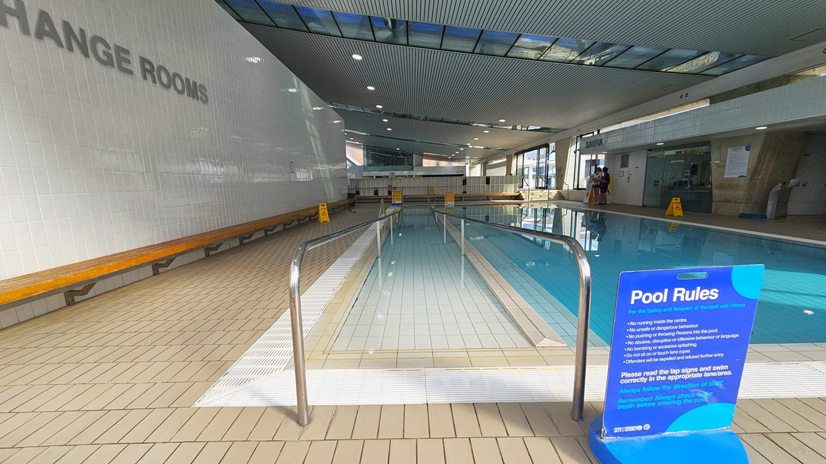 The Program Pool is equipped with ramp entry and handrails. Freestanding banners displaying Pool Rules and safety warnings are affixed to the pool deck. A long wooden bench is positioned against the left wall. The ground floor is tiled with small bricks, and the ceiling features a white porous design with circular LED lights, reflecting the pool below. To the left is a white brick wall with a sign indicating "Change rooms." The Program Pool is equipped with ramp entry and handrails. Freestanding banners displaying Pool Rules and safety warnings are affixed to the pool deck. A long wooden bench is positioned against the left wall. The ground floor is tiled with small bricks, and the ceiling features a white porous design with circular LED lights, reflecting the pool below. To the left is a white brick wall with a sign indicating "Change rooms."
