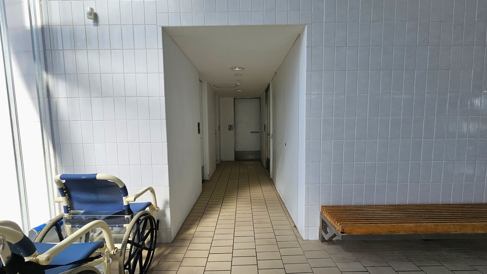 Entrance to the accessible bathrooms. Two aquatic wheelchairs are positioned on the left, and a long wooden bench is on the right. The wall is made of small bricks. The corridor leading to the bathrooms has cream floors, white walls, and white ceilings with circular LED lights. Entrance to the accessible bathrooms. Two aquatic wheelchairs are positioned on the left, and a long wooden bench is on the right. The wall is made of small bricks. The corridor leading to the bathrooms has cream floors, white walls, and white ceilings with circular LED lights.