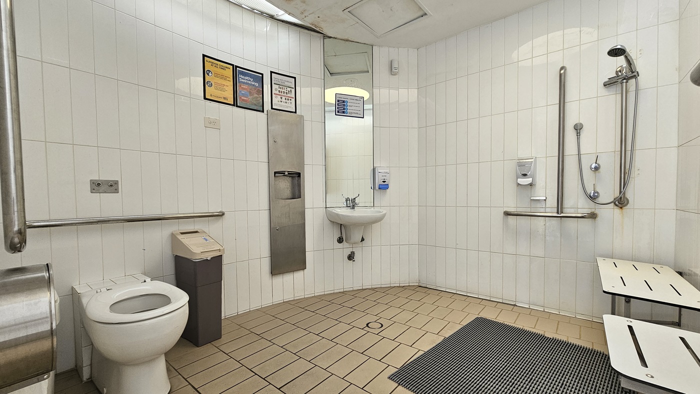 Within the accessible bathroom, a toilet bowl with hand grabs is situated on the left, accompanied by a tissue dispenser and a rubbish bin. Adjacent to it on the left wall are a wash basin, tissue dispenser, mirrors, and soap dispenser. On the right, there is a shower with handrails and a foldable bench is placed on the right wall. Banners are affixed to the upper left wall. The walls are adorned with white tiles, and the floor is covered in brown tiles with a non-slippery mat. Within the accessible bathroom, a toilet bowl with hand grabs is situated on the left, accompanied by a tissue dispenser and a rubbish bin. Adjacent to it on the left wall are a wash basin, tissue dispenser, mirrors, and soap dispenser. On the right, there is a shower with handrails and a foldable bench is placed on the right wall. Banners are affixed to the upper left wall. The walls are adorned with white tiles, and the floor is covered in brown tiles with a non-slippery mat.