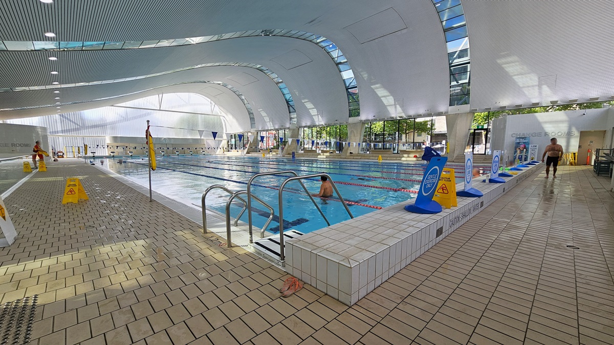 The Lap pool features steps with handrails, floating ropes, and blue and white buntings running across the pool. Freestanding banners line the raised pool deck. The change room is on the right, and safety warning signs and truncated domes are on the left pool deck. Some guests are using the pool. The ground floor is tiled with small bricks, and the wave-shaped ceiling displays a white porous design with circular LED lights, reflecting the pool below. The Lap pool features steps with handrails, floating ropes, and blue and white buntings running across the pool. Freestanding banners line the raised pool deck. The change room is on the right, and safety warning signs and truncated domes are on the left pool deck. Some guests are using the pool. The ground floor is tiled with small bricks, and the wave-shaped ceiling displays a white porous design with circular LED lights, reflecting the pool below.