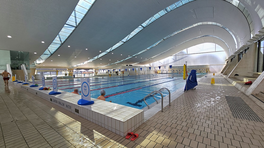 The Lap pool has steps with handrails, equipped with truncated domes. Floating ropes and blue and white buntings run across the pool. Freestanding banners line the raised pool deck on the left. Some guests are using the pool. The floor is tiled with small bricks, and the wave-shaped ceiling features a white porous design with circular LED lights, reflecting the pool below. The Lap pool has steps with handrails, equipped with truncated domes. Floating ropes and blue and white buntings run across the pool. Freestanding banners line the raised pool deck on the left. Some guests are using the pool. The floor is tiled with small bricks, and the wave-shaped ceiling features a white porous design with circular LED lights, reflecting the pool below.