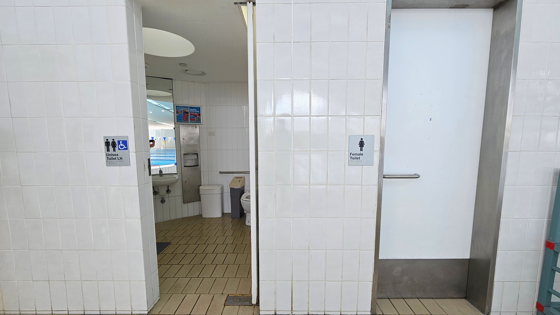 The left door leads to an all-gender and accessible toilet. Inside, the toilet bowl is positioned against the wall, accompanied by rubbish bins, mirrors, a washbasin, and a tissue dispenser. The floor is covered in brown tiles, the walls are adorned with white tiles, and the ceiling features circular LED lights. The left door designates a female toilet. The left door leads to an all-gender and accessible toilet. Inside, the toilet bowl is positioned against the wall, accompanied by rubbish bins, mirrors, a washbasin, and a tissue dispenser. The floor is covered in brown tiles, the walls are adorned with white tiles, and the ceiling features circular LED lights. The left door designates a female toilet.