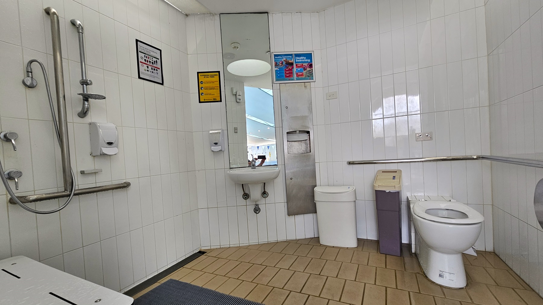 Within the accessible bathroom, the toilet bowl is against the right wall with accompanying rubbish bins. Mirrors, a washbasin, and a tissue dispenser are mounted on the left wall, along with showers featuring handgrabs. Adjacent to these fixtures is a foldable bench. The floor is covered in brown tiles with a non-slippery mat, and the walls are adorned with white tiles. The ceiling is equipped with circular LED lights. The left door designates a female toilet. Within the accessible bathroom, the toilet bowl is against the right wall with accompanying rubbish bins. Mirrors, a washbasin, and a tissue dispenser are mounted on the left wall, along with showers featuring handgrabs. Adjacent to these fixtures is a foldable bench. The floor is covered in brown tiles with a non-slippery mat, and the walls are adorned with white tiles. The ceiling is equipped with circular LED lights. The left door designates a female toilet.