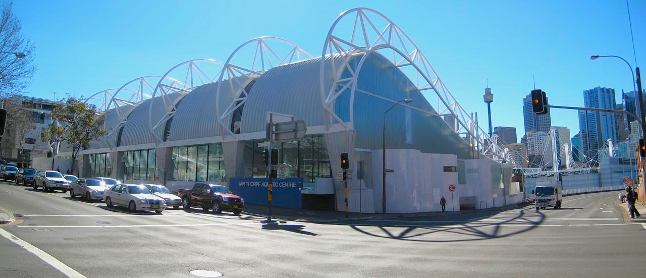 At a road intersection stands a modern commercial building with a unique blue corrugated wave roofing design. White metal structures complement the roofing contours. A blue wall displaying the facility sign is on the left. To the right, a stop light is visible, with cars stopped on the left side. Further back, tall commercial buildings can be seen. At a road intersection stands a modern commercial building with a unique blue corrugated wave roofing design. White metal structures complement the roofing contours. A blue wall displaying the facility sign is on the left. To the right, a stop light is visible, with cars stopped on the left side. Further back, tall commercial buildings can be seen.