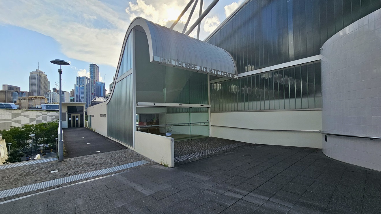 The building showcases a distinctive corrugated wave roofing design in blue. The left wall features glass structures, and a glass door entry is on the left. The facility name is written on top of the wave-shaped roofing. The entrance has an automatic glass sliding door with truncated domes. To the left is a pathway leading to another entrance, overlooking the section below. Tall light posts are present. Further ahead, tall commercial buildings come into view. The building showcases a distinctive corrugated wave roofing design in blue. The left wall features glass structures, and a glass door entry is on the left. The facility name is written on top of the wave-shaped roofing. The entrance has an automatic glass sliding door with truncated domes. To the left is a pathway leading to another entrance, overlooking the section below. Tall light posts are present. Further ahead, tall commercial buildings come into view.