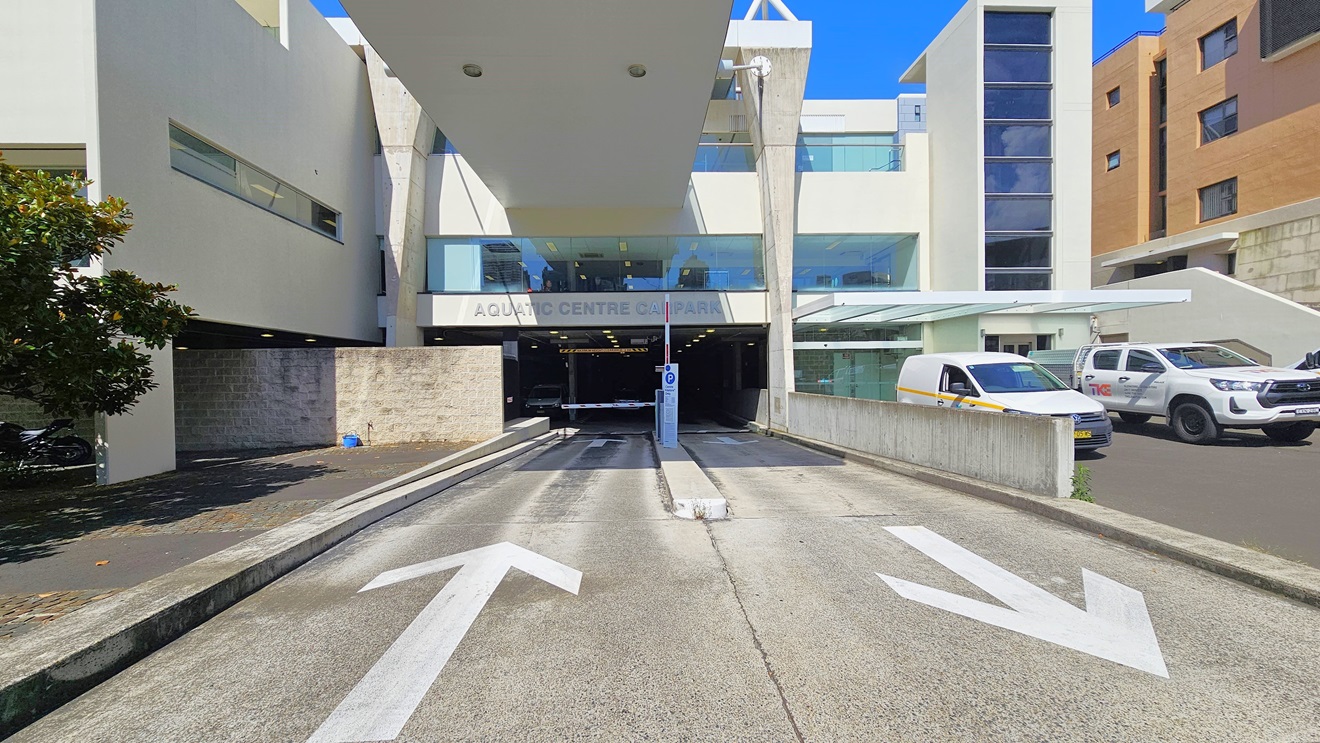 The two-way road provides access to and from the underground car park. The facility name is displayed before the entrance, separated by a traffic median. Limited parking is available on the right, with two cars currently parked. A brown facility building is situated on the right, and a tree is visible in the concrete area on the left. The two-way road provides access to and from the underground car park. The facility name is displayed before the entrance, separated by a traffic median. Limited parking is available on the right, with two cars currently parked. A brown facility building is situated on the right, and a tree is visible in the concrete area on the left.