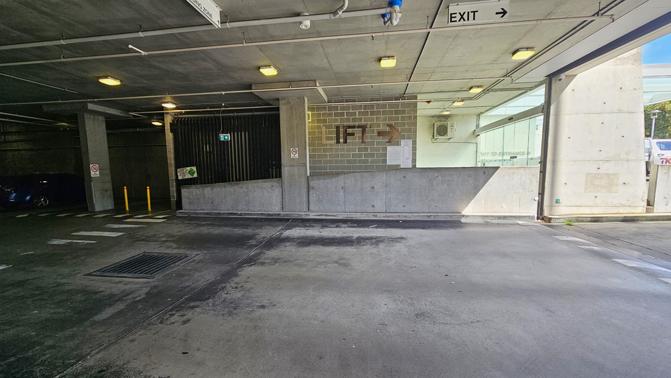 The underground parking includes a pedestrian lane on the left, marked by yellow bollards. The middle brick wall has a sign indicating the lift direction. The low ceiling features directional signs, LED lights, and exposed pipes. Safety signs are affixed to the wall, and a visible water drainage system is present. The underground parking includes a pedestrian lane on the left, marked by yellow bollards. The middle brick wall has a sign indicating the lift direction. The low ceiling features directional signs, LED lights, and exposed pipes. Safety signs are affixed to the wall, and a visible water drainage system is present.
