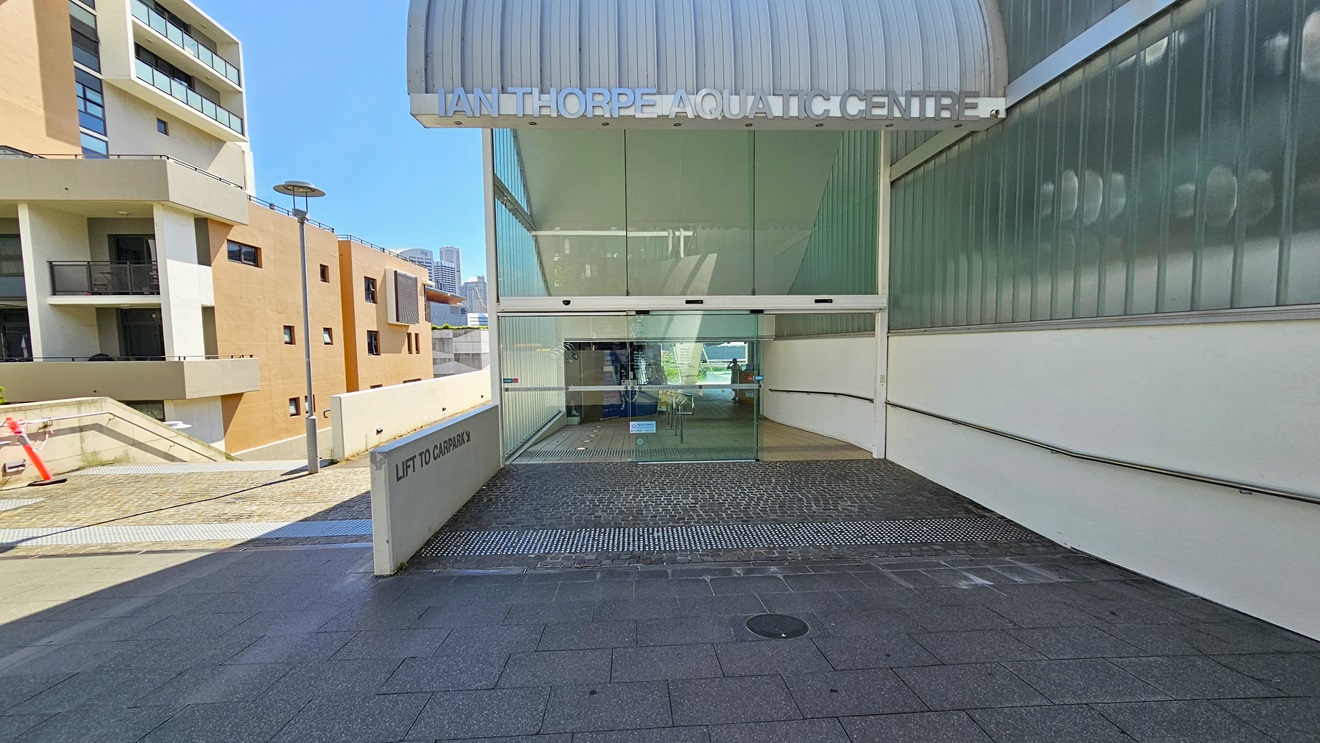 The facility entrance is through an automatic glass sliding door, with truncated domes on the floor. The right wall features a glass structure, and the facility name is written on top of the wave-shaped roofing. To the left is a multi-story cream building, with a direction leading to the car park visible on the left. The facility entrance is through an automatic glass sliding door, with truncated domes on the floor. The right wall features a glass structure, and the facility name is written on top of the wave-shaped roofing. To the left is a multi-story cream building, with a direction leading to the car park visible on the left.
