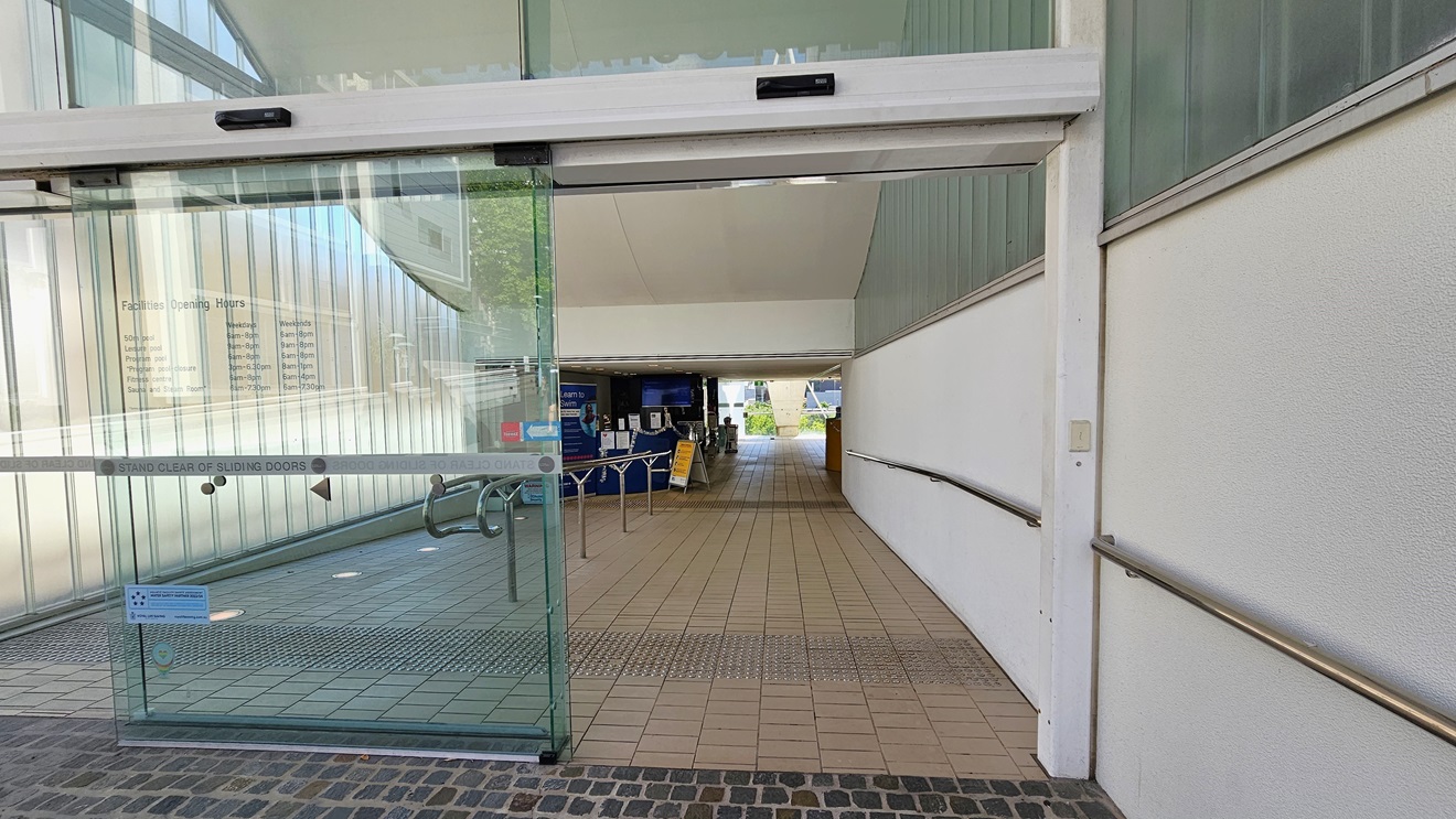 The automatic glass sliding door displays the facility sticker. Truncated domes are installed on the floor. The lobby path has a gentle descent, with handrails positioned in the middle and on the right. Moving ahead, the reception is visible. The floor is covered with grey tiles. The automatic glass sliding door displays the facility sticker. Truncated domes are installed on the floor. The lobby path has a gentle descent, with handrails positioned in the middle and on the right. Moving ahead, the reception is visible. The floor is covered with grey tiles.