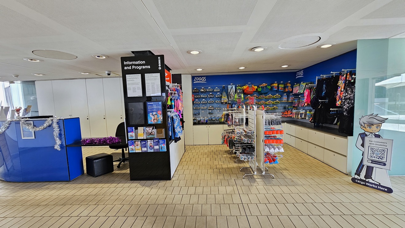 In the center is the retail section showcasing goggles, swimwear, and swimming accessories. On the left, a blue desk adorned with Christmas garland is dedicated to the Learn to Swim program, with brochures displayed in front of the black display rack. The floor is tiled with small bricks, and the ceiling features a white porous design with circular LED lights. In the center is the retail section showcasing goggles, swimwear, and swimming accessories. On the left, a blue desk adorned with Christmas garland is dedicated to the Learn to Swim program, with brochures displayed in front of the black display rack. The floor is tiled with small bricks, and the ceiling features a white porous design with circular LED lights.