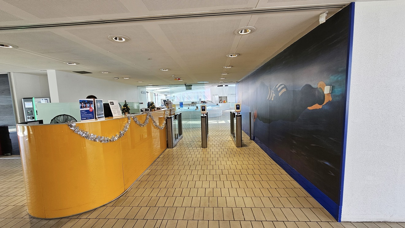 In the center stands the reception desk, featuring a caramel-brown surface embellished with Christmas garlands. To its right, a glass turnstile is in place for controlled entry. On the left, there is a spacious area with foam seating. The right wall showcases a life-sized painting of Ian Thorpe swimming underwater. The floor is tiled with small bricks, and the ceiling exhibits a white porous design with circular LED lights. In the center stands the reception desk, featuring a caramel-brown surface embellished with Christmas garlands. To its right, a glass turnstile is in place for controlled entry. On the left, there is a spacious area with foam seating. The right wall showcases a life-sized painting of Ian Thorpe swimming underwater. The floor is tiled with small bricks, and the ceiling exhibits a white porous design with circular LED lights.