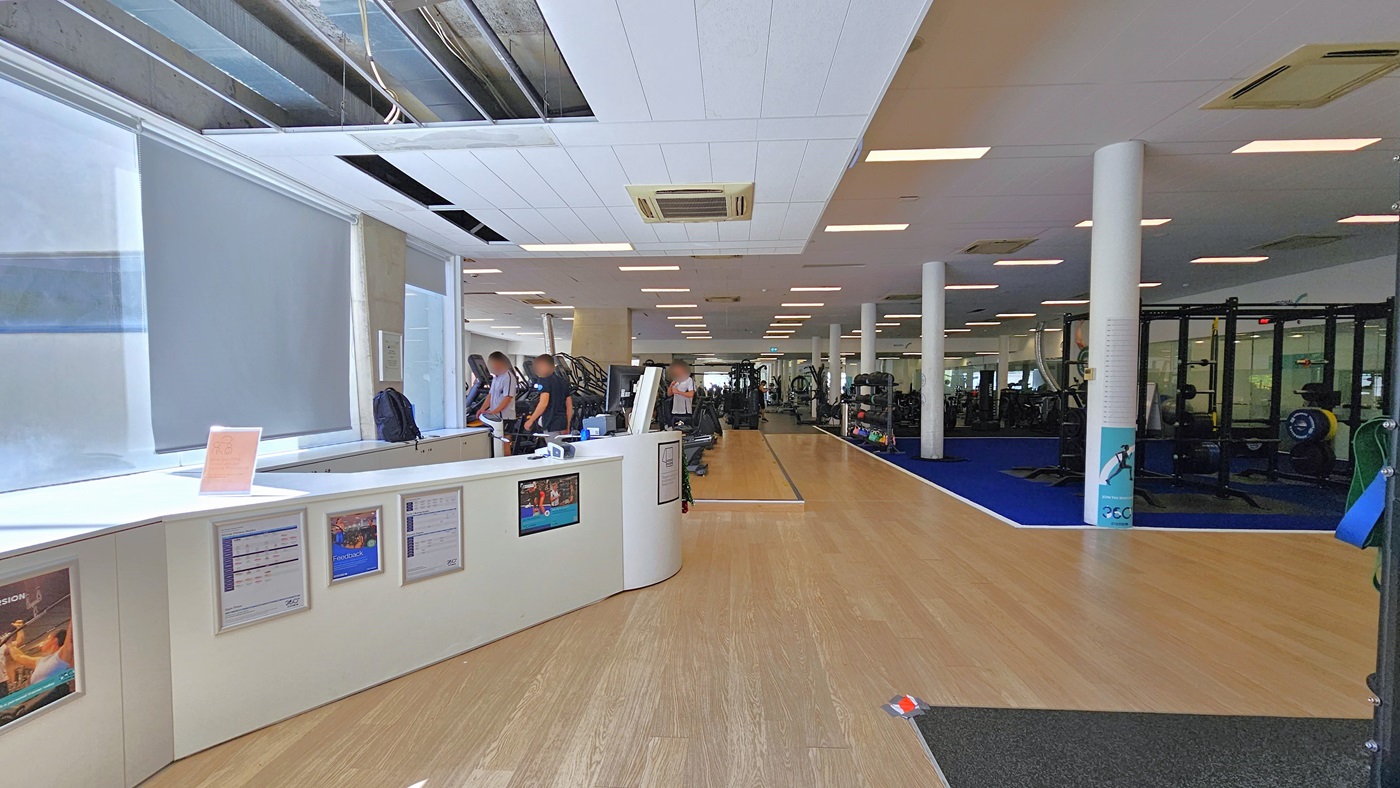 Access to the gym is through an inward-opening manual glass door. Inside, turnstiles equipped with swiping machines for access are positioned at the entry. The floor inside features a combination of black and white tiles, and the ceiling is white with centralized air conditioning. Outside, to the left, there is a lift. Access to the gym is through an inward-opening manual glass door. Inside, turnstiles equipped with swiping machines for access are positioned at the entry. The floor inside features a combination of black and white tiles, and the ceiling is white with centralized air conditioning. Outside, to the left, there is a lift.