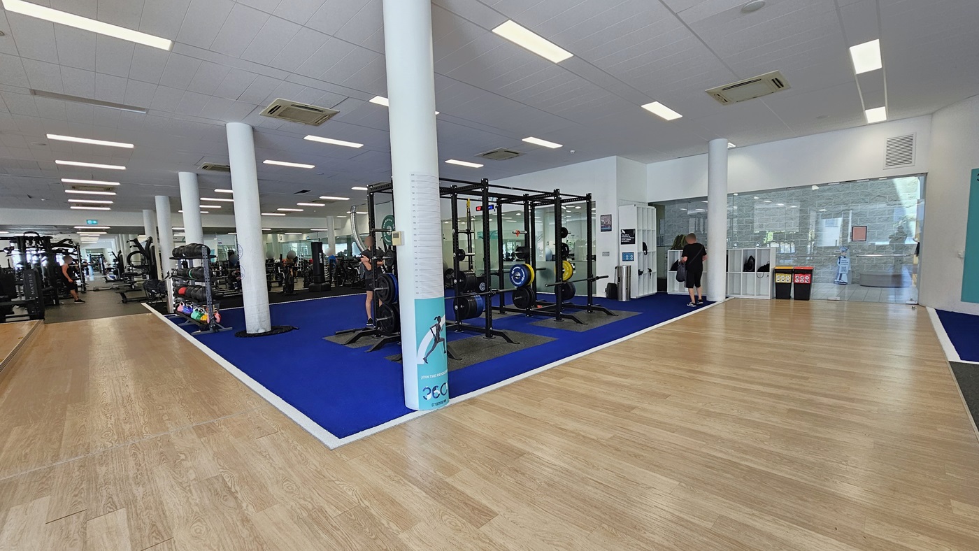 A royal blue rubber mat in the middle accommodates Smith Machines. Further in, stability balls, free weights, and additional machines are on display. To the right, there are white storage racks for belongings, and adjacent to them are two rubbish bins. The floor transitions from timber to blue rubber mats. The ceiling is white with a porous design, featuring LED lights and centralized air conditioning. A royal blue rubber mat in the middle accommodates Smith Machines. Further in, stability balls, free weights, and additional machines are on display. To the right, there are white storage racks for belongings, and adjacent to them are two rubbish bins. The floor transitions from timber to blue rubber mats. The ceiling is white with a porous design, featuring LED lights and centralized air conditioning.