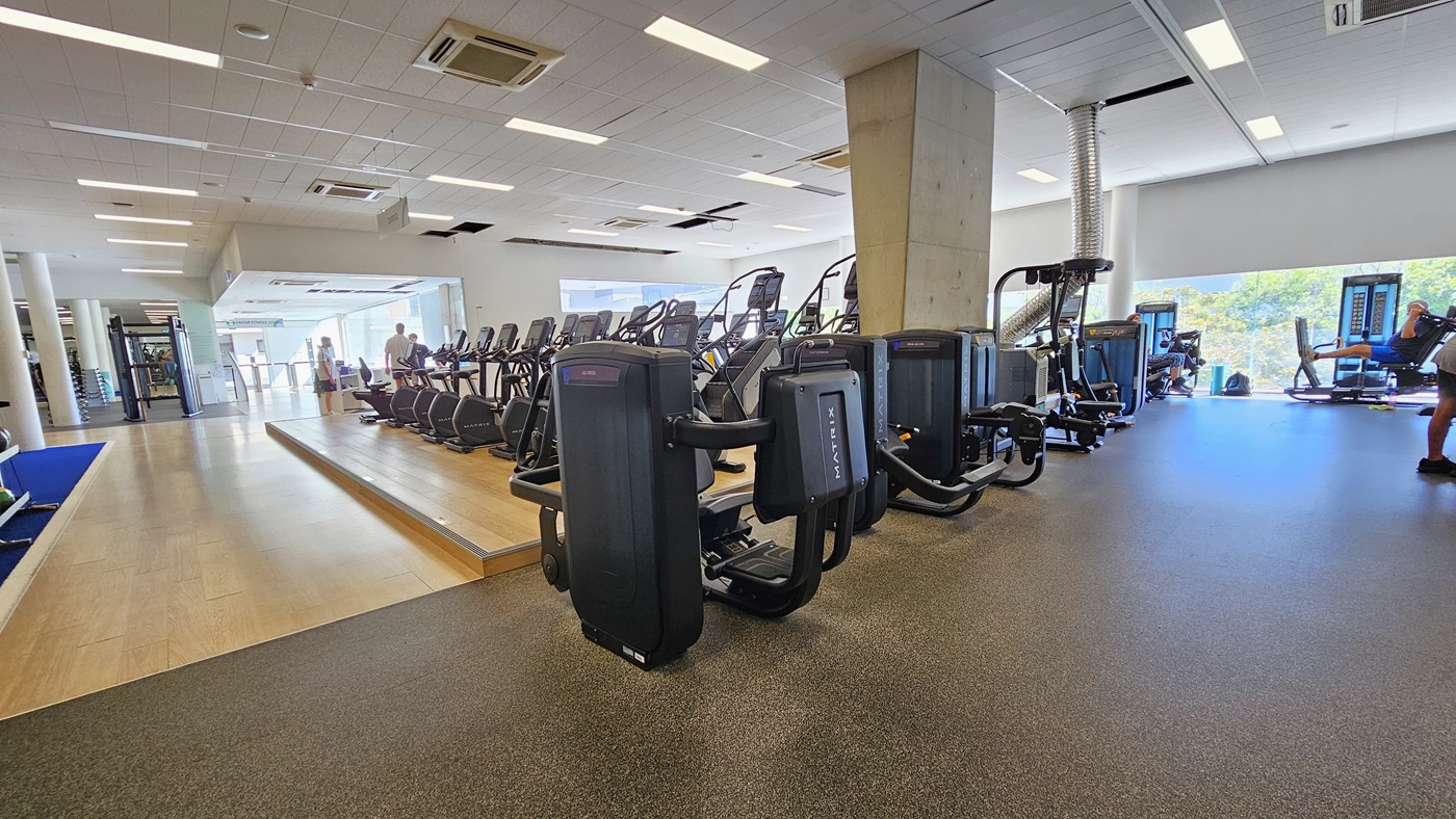 In the middle, a slightly elevated platform accommodates a lineup of cardio machines. To the left, a royal blue mat hosts stability and free weights, while further along, Smith Machines are positioned. To the right, more machines are arranged. The floor transitions from a blue rubber mat to timber in the middle and a black rubber mat on the right. The ceiling is white with a porous design, equipped with LED lights and centralized air conditioning. In the middle, a slightly elevated platform accommodates a lineup of cardio machines. To the left, a royal blue mat hosts stability and free weights, while further along, Smith Machines are positioned. To the right, more machines are arranged. The floor transitions from a blue rubber mat to timber in the middle and a black rubber mat on the right. The ceiling is white with a porous design, equipped with LED lights and centralized air conditioning.