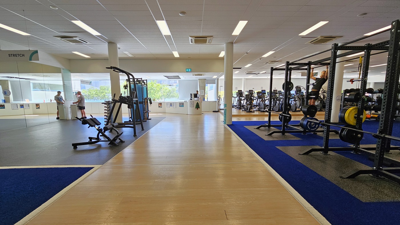 On the left is a glass door entrance with stretch machines positioned. The reception desk is in the middle. To the right, Smith machines and free weights are lined up, directly adjacent to cardio machines. The floor transitions from a blue rubber mat to a timber and a black rubber mat on the right. The ceiling is white with a porous design, featuring LED lights and centralized air conditioning. Few gym users are visible. On the left is a glass door entrance with stretch machines positioned. The reception desk is in the middle. To the right, Smith machines and free weights are lined up, directly adjacent to cardio machines. The floor transitions from a blue rubber mat to a timber and a black rubber mat on the right. The ceiling is white with a porous design, featuring LED lights and centralized air conditioning. Few gym users are visible.