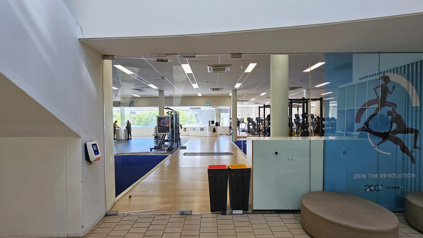The manual inward-opening glass door serves as an alternate entrance to the gym. A swiping machine is positioned on the left for members to swipe their access cards. To the right, round cushion seatings are available. A large sports-related poster is affixed to the left glass wall. Inside, the gym's timber floor and blue mat are visible, with machines strategically positioned. Two rubbish bins are placed at the entrance. Gym users are also visible. The manual inward-opening glass door serves as an alternate entrance to the gym. A swiping machine is positioned on the left for members to swipe their access cards. To the right, round cushion seatings are available. A large sports-related poster is affixed to the left glass wall. Inside, the gym's timber floor and blue mat are visible, with machines strategically positioned. Two rubbish bins are placed at the entrance. Gym users are also visible.