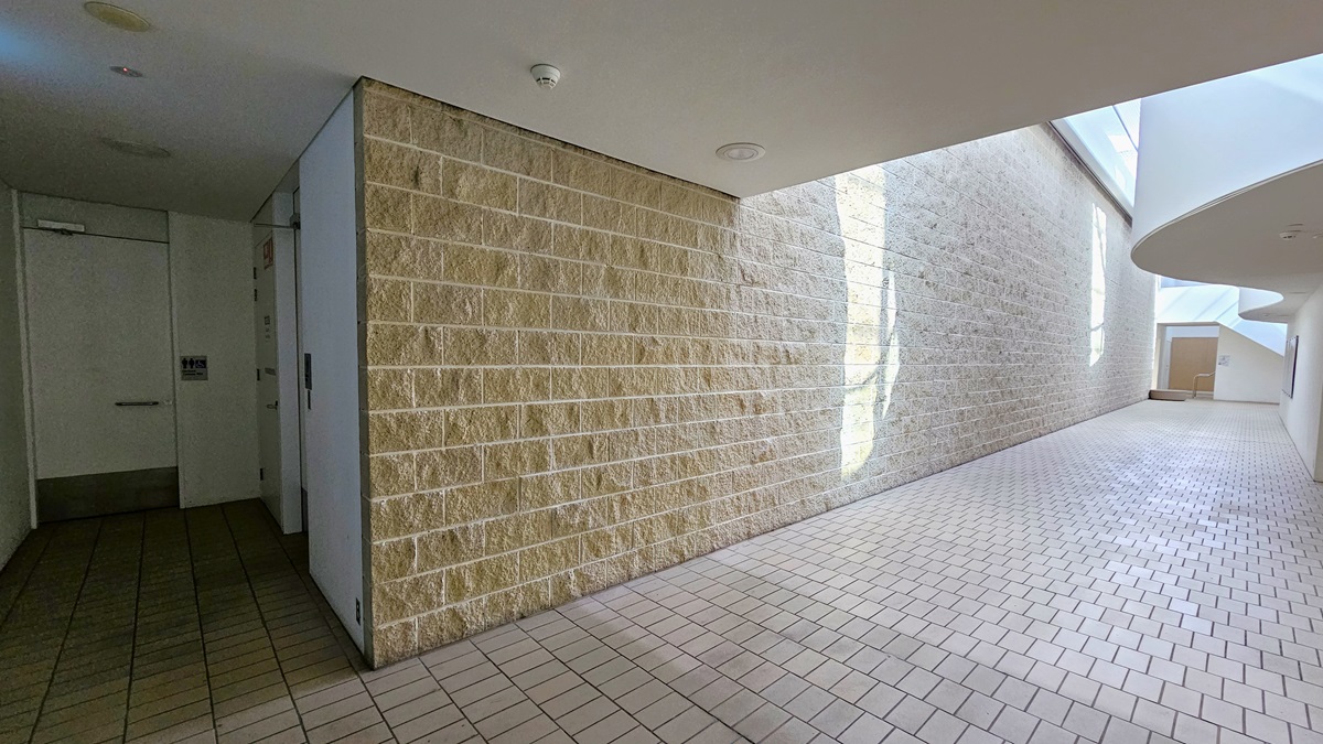 To the right is a corridor leading to the accessible bathrooms. The walls are constructed with brown bricks, and the floor is covered in brown tiles. On the left, there are two accessible bathrooms, each marked with signage indicating gender access. The ceiling is entirely white, featuring circular LED lights. To the right is a corridor leading to the accessible bathrooms. The walls are constructed with brown bricks, and the floor is covered in brown tiles. On the left, there are two accessible bathrooms, each marked with signage indicating gender access. The ceiling is entirely white, featuring circular LED lights.