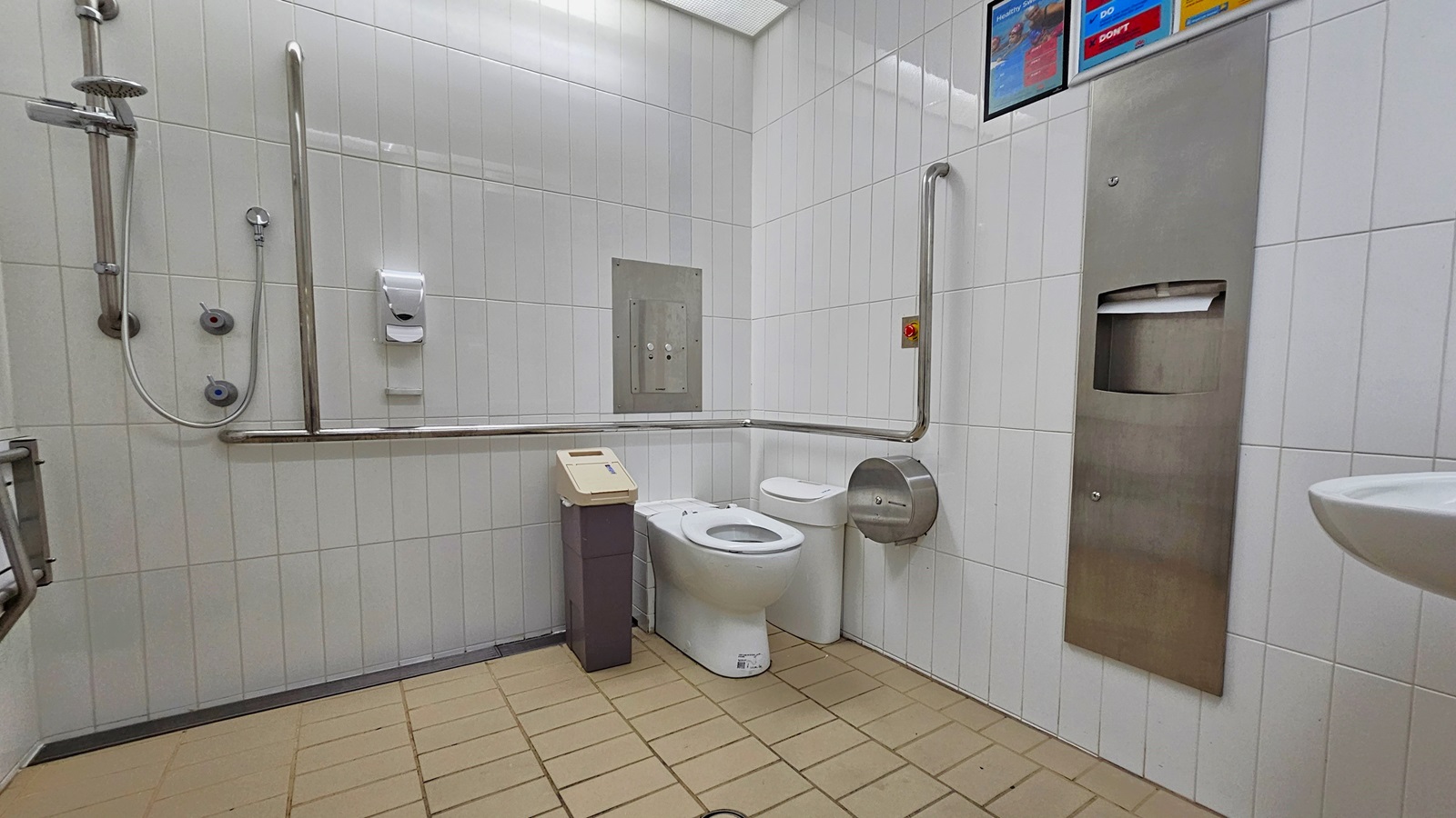 Inside the accessible bathroom. The toilet bowl, equipped with a handrail, is positioned in the right corner. Rubbish bins and a tissue dispenser are placed beside it. To the right, there is another tissue dispenser and a washbasin mounted on the wall. On the left, a shower with handrails is present, and a foldable bench is mounted on the left wall. The walls are adorned with white tiles, featuring posters affixed on top. The floor is covered in cream-coloured tiles. Inside the accessible bathroom. The toilet bowl, equipped with a handrail, is positioned in the right corner. Rubbish bins and a tissue dispenser are placed beside it. To the right, there is another tissue dispenser and a washbasin mounted on the wall. On the left, a shower with handrails is present, and a foldable bench is mounted on the left wall. The walls are adorned with white tiles, featuring posters affixed on top. The floor is covered in cream-coloured tiles.