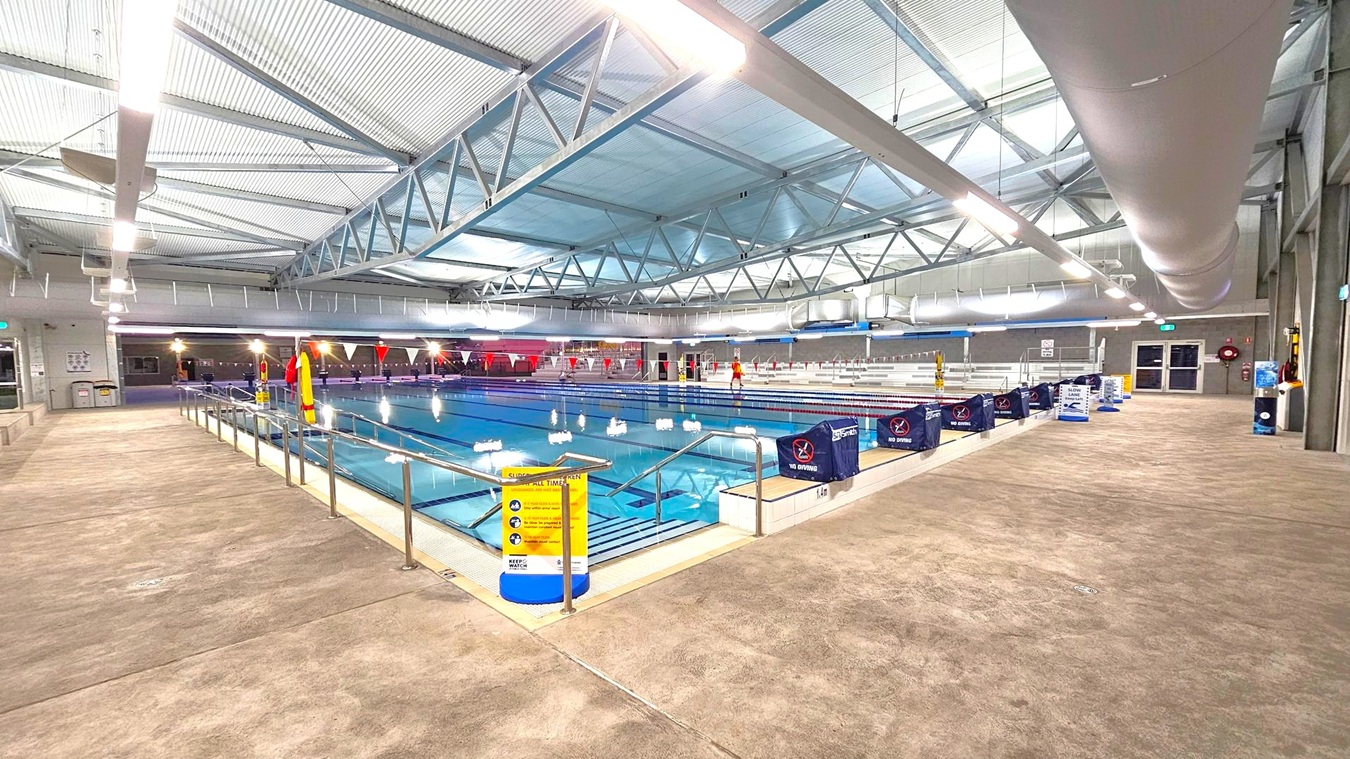 Indoor swimming pool with multiple lanes, surrounded by a concrete floor. On the left, a railing runs alongside the pool with a yellow safety sign. The right side features a series of blue "No Diving" signs. In the background, there are tiered seating areas. The ceiling is high with exposed metal beams and large ventilation ducts. Bright overhead lights illuminate the space. Indoor swimming pool with multiple lanes, surrounded by a concrete floor. On the left, a railing runs alongside the pool with a yellow safety sign. The right side features a series of blue "No Diving" signs. In the background, there are tiered seating areas. The ceiling is high with exposed metal beams and large ventilation ducts. Bright overhead lights illuminate the space.