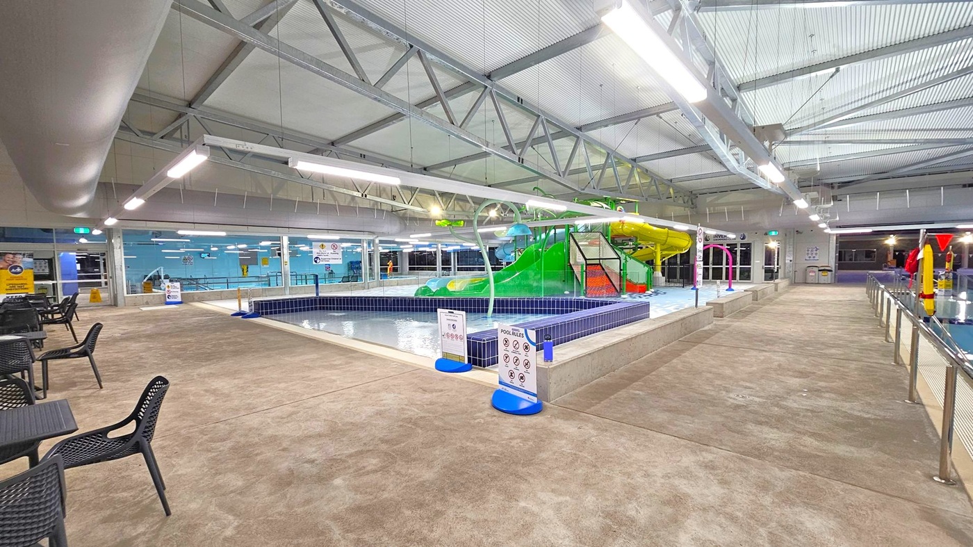 Indoor swimming pool area with a children's play section in the centre featuring a green and yellow slide. The pool is surrounded by a tiled edge with pool rules signs. On the left, there are black chairs and tables on a beige floor. The ceiling has exposed metal beams and bright overhead lights. On the right, a railing separates the walkway from the pool area. The background shows blue walls with windows and additional signage. Indoor swimming pool area with a children's play section in the centre featuring a green and yellow slide. The pool is surrounded by a tiled edge with pool rules signs. On the left, there are black chairs and tables on a beige floor. The ceiling has exposed metal beams and bright overhead lights. On the right, a railing separates the walkway from the pool area. The background shows blue walls with windows and additional signage.