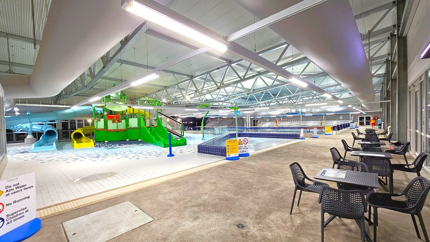 Indoor swimming pool area with a children's play section on the left, featuring colourful slides in blue, yellow, and green. The middle has a shallow pool with a tiled edge. On the right, there are several black tables and chairs on a concrete floor. The ceiling is high with exposed beams and large ventilation ducts. Bright overhead lights illuminate the space. Signs with safety instructions are visible near the pool. Indoor swimming pool area with a children's play section on the left, featuring colourful slides in blue, yellow, and green. The middle has a shallow pool with a tiled edge. On the right, there are several black tables and chairs on a concrete floor. The ceiling is high with exposed beams and large ventilation ducts. Bright overhead lights illuminate the space. Signs with safety instructions are visible near the pool.