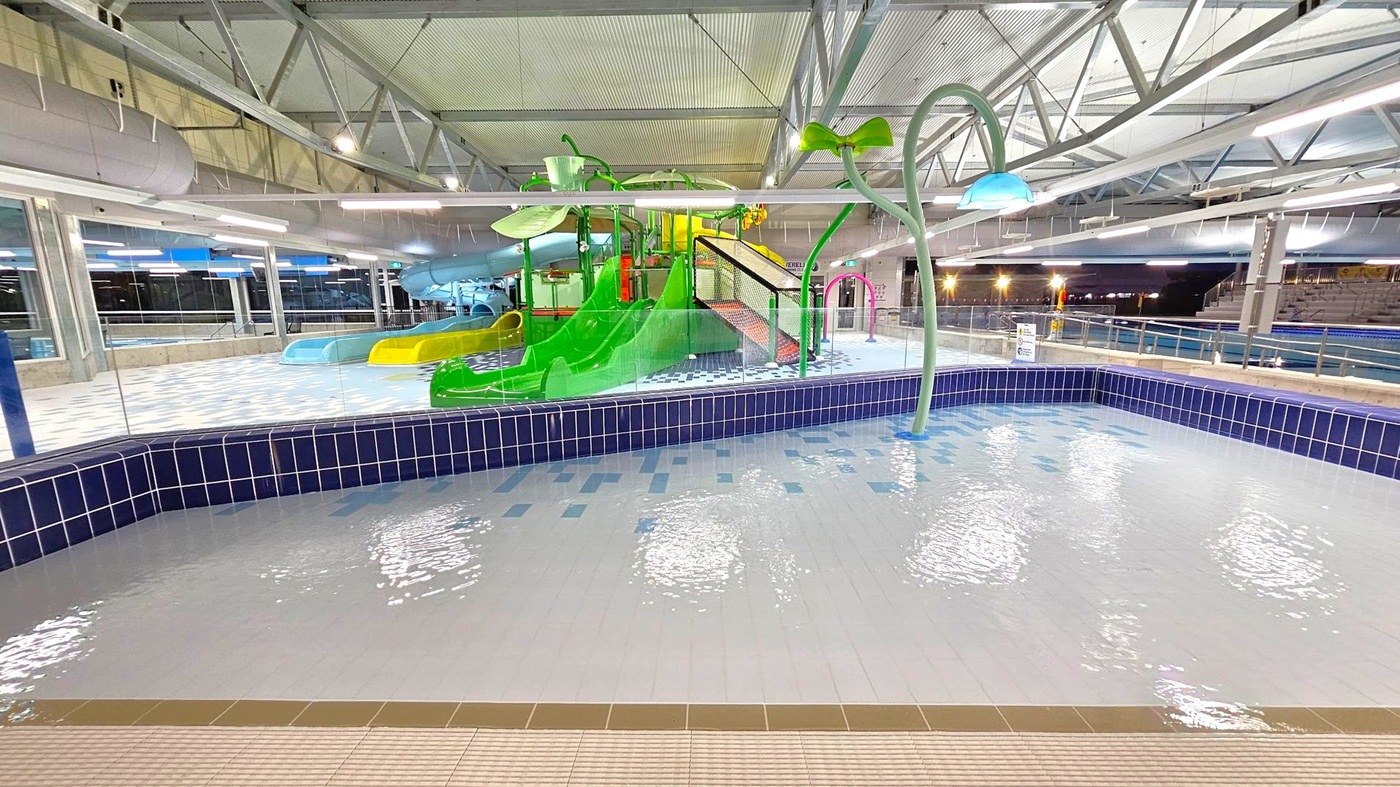 An indoor water play area with a shallow pool in the foreground, featuring light blue and white tiles. In the middle, a colourful play structure with green, yellow, and blue slides is visible. A large green and blue water feature resembling a flower stands to the right. The background shows a spacious area with metal beams and large windows, providing a view of the outside. The ceiling is high with exposed metal beams and lighting fixtures. An indoor water play area with a shallow pool in the foreground, featuring light blue and white tiles. In the middle, a colourful play structure with green, yellow, and blue slides is visible. A large green and blue water feature resembling a flower stands to the right. The background shows a spacious area with metal beams and large windows, providing a view of the outside. The ceiling is high with exposed metal beams and lighting fixtures.