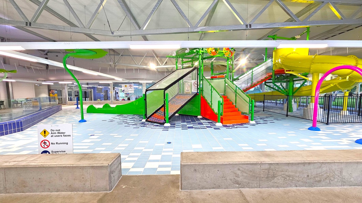 Indoor water play area with a colourful play structure in the centre. The structure has green and orange steps leading up to a platform with a yellow spiral slide. The floor is tiled in shades of blue and white. A sign in the foreground warns against aiming water at faces and running. Overhead, the ceiling features exposed metal beams and fluorescent lighting. On the right, a purple water feature is visible, and a black fence encloses the slide area. Indoor water play area with a colourful play structure in the centre. The structure has green and orange steps leading up to a platform with a yellow spiral slide. The floor is tiled in shades of blue and white. A sign in the foreground warns against aiming water at faces and running. Overhead, the ceiling features exposed metal beams and fluorescent lighting. On the right, a purple water feature is visible, and a black fence encloses the slide area.