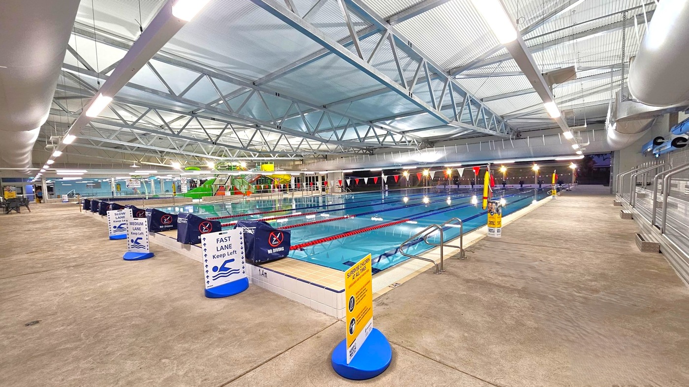 Indoor swimming pool with multiple lanes, each marked by signs for slow, medium, and fast swimmers. The pool is surrounded by a concrete floor with a few wet patches. On the left, there are signs and barriers, while on the right, a metal railing and steps lead into the pool. The ceiling features exposed metal beams and bright overhead lights. In the background, there are colourful water slides and additional pool facilities. Indoor swimming pool with multiple lanes, each marked by signs for slow, medium, and fast swimmers. The pool is surrounded by a concrete floor with a few wet patches. On the left, there are signs and barriers, while on the right, a metal railing and steps lead into the pool. The ceiling features exposed metal beams and bright overhead lights. In the background, there are colourful water slides and additional pool facilities.