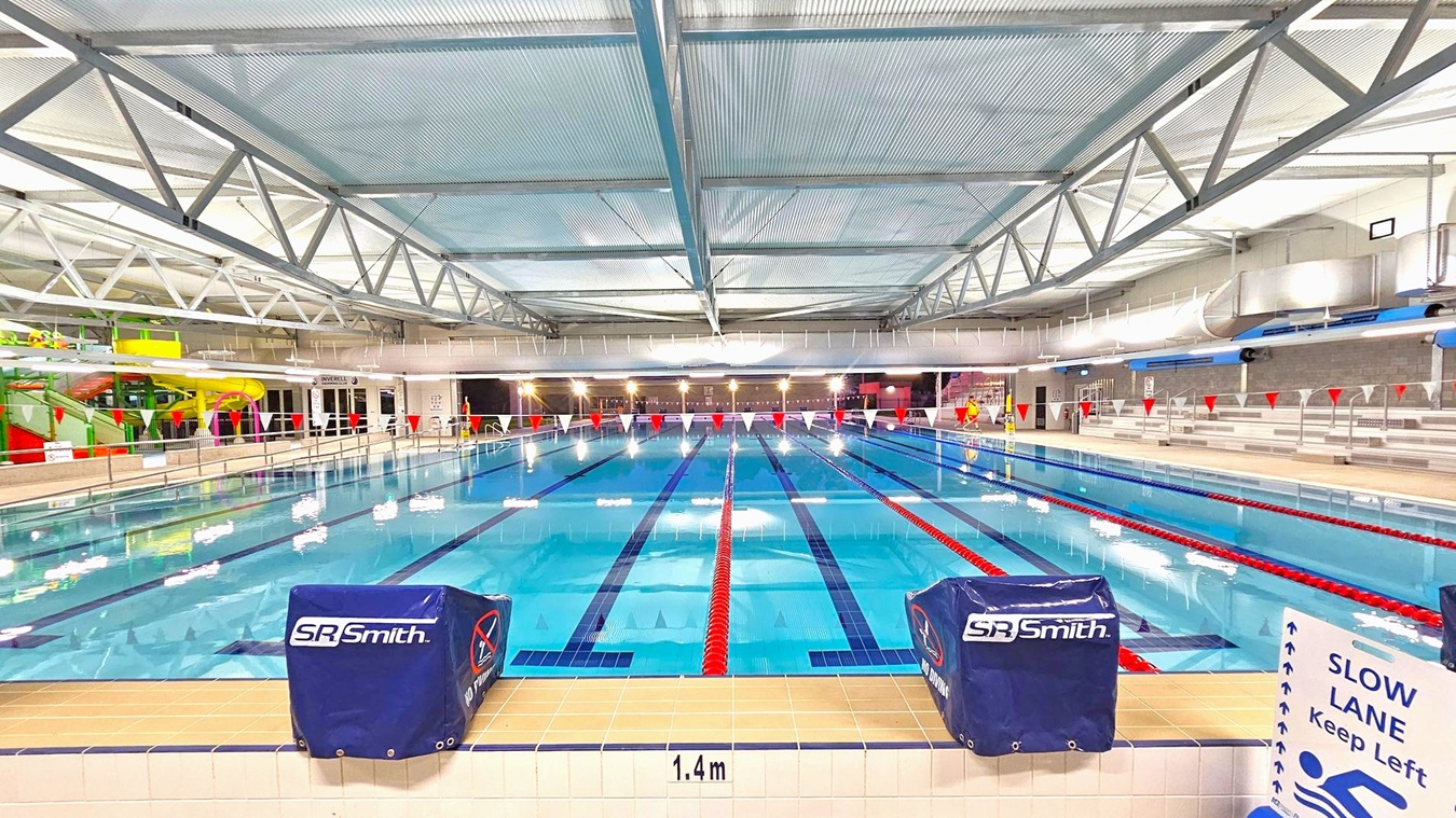 Indoor swimming pool with multiple lanes, marked by red and white lane dividers. The pool is surrounded by tiled flooring with a depth marker of 1.4m at the edge. On the left, there is a colourful water slide structure. On the right, tiered seating is visible. The ceiling features metal trusses and translucent panels. A sign on the right indicates a slow lane. The pool area is well-lit with overhead lights. Indoor swimming pool with multiple lanes, marked by red and white lane dividers. The pool is surrounded by tiled flooring with a depth marker of 1.4m at the edge. On the left, there is a colourful water slide structure. On the right, tiered seating is visible. The ceiling features metal trusses and translucent panels. A sign on the right indicates a slow lane. The pool area is well-lit with overhead lights.