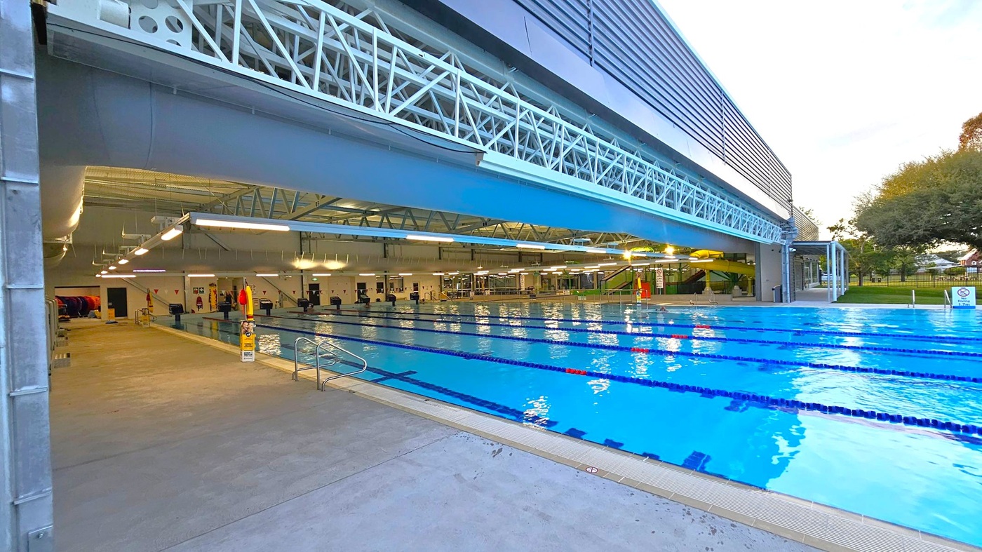 An indoor swimming pool with multiple lanes stretches from the foreground to the background. On the left, a concrete walkway runs alongside the pool, with safety signs and a ladder. Overhead, a metal truss structure supports the ceiling, which is equipped with fluorescent lights. In the background, there are changing rooms and a green slide. To the right, the pool extends outdoors, bordered by a grassy area with trees. The ceiling is industrial with exposed beams. An indoor swimming pool with multiple lanes stretches from the foreground to the background. On the left, a concrete walkway runs alongside the pool, with safety signs and a ladder. Overhead, a metal truss structure supports the ceiling, which is equipped with fluorescent lights. In the background, there are changing rooms and a green slide. To the right, the pool extends outdoors, bordered by a grassy area with trees. The ceiling is industrial with exposed beams.