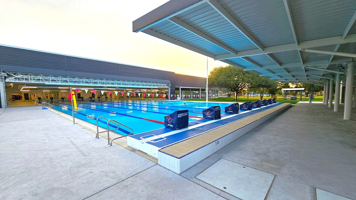 An outdoor swimming pool with multiple lanes, each marked by red and white lane dividers. On the right, a row of starting blocks is positioned along the pool's edge. A covered walkway with a metal roof and supporting columns runs parallel to the pool. On the left, a building with large windows and a dark exterior is visible. In the background, trees and a grassy area can be seen. The floor is paved with concrete tiles. An outdoor swimming pool with multiple lanes, each marked by red and white lane dividers. On the right, a row of starting blocks is positioned along the pool's edge. A covered walkway with a metal roof and supporting columns runs parallel to the pool. On the left, a building with large windows and a dark exterior is visible. In the background, trees and a grassy area can be seen. The floor is paved with concrete tiles.