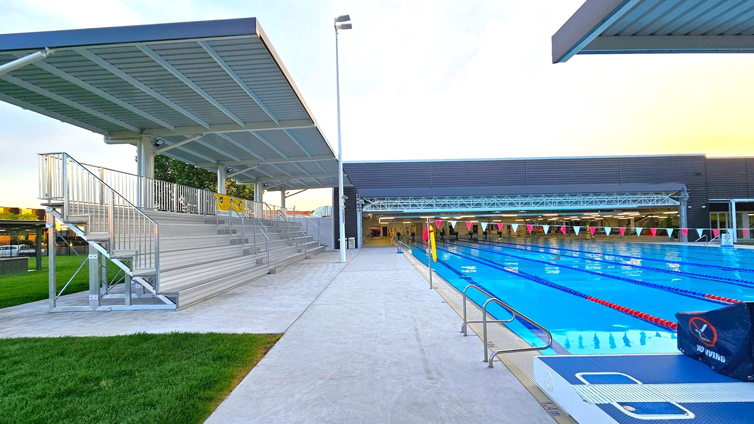 On the left, there are metal bleachers with a corrugated metal roof, supported by metal beams. A set of stairs leads up to the seating area. The ground is a mix of concrete and grass. On the right, a swimming pool with multiple lanes is visible, marked by lane dividers and triangular flags overhead. A "No Diving" sign is placed near the pool's edge. In the background, a building with large windows and a flat roof is visible, with a covered walkway connecting to it. On the left, there are metal bleachers with a corrugated metal roof, supported by metal beams. A set of stairs leads up to the seating area. The ground is a mix of concrete and grass. On the right, a swimming pool with multiple lanes is visible, marked by lane dividers and triangular flags overhead. A "No Diving" sign is placed near the pool's edge. In the background, a building with large windows and a flat roof is visible, with a covered walkway connecting to it.
