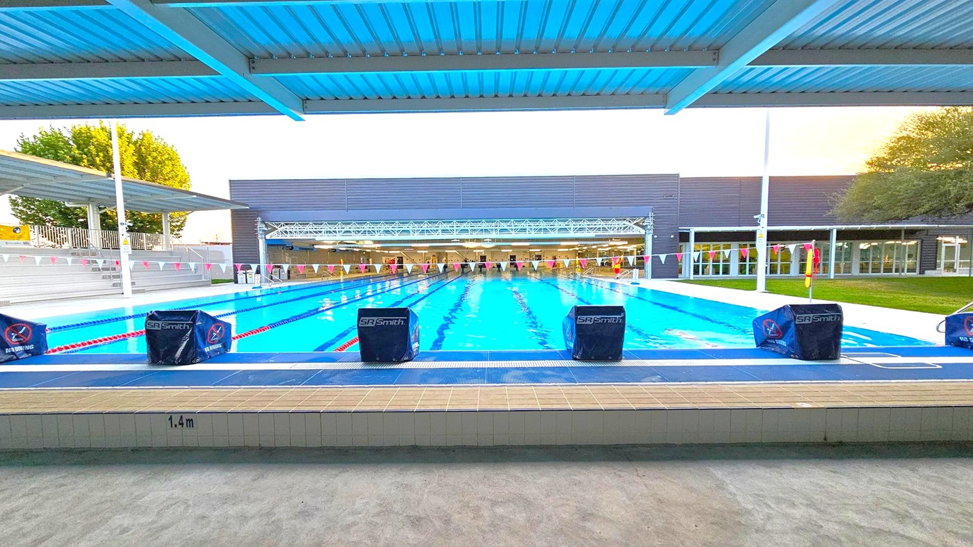 An outdoor swimming pool with clear blue water, marked by lane dividers. Starting blocks are positioned at the pool's edge. A modern building with large windows is in the background, and a tree is visible to the left. The pool area is covered by a metal roof, and the floor is tiled. A grassy area is on the right, and a row of flags hangs above the pool. An outdoor swimming pool with clear blue water, marked by lane dividers. Starting blocks are positioned at the pool's edge. A modern building with large windows is in the background, and a tree is visible to the left. The pool area is covered by a metal roof, and the floor is tiled. A grassy area is on the right, and a row of flags hangs above the pool.