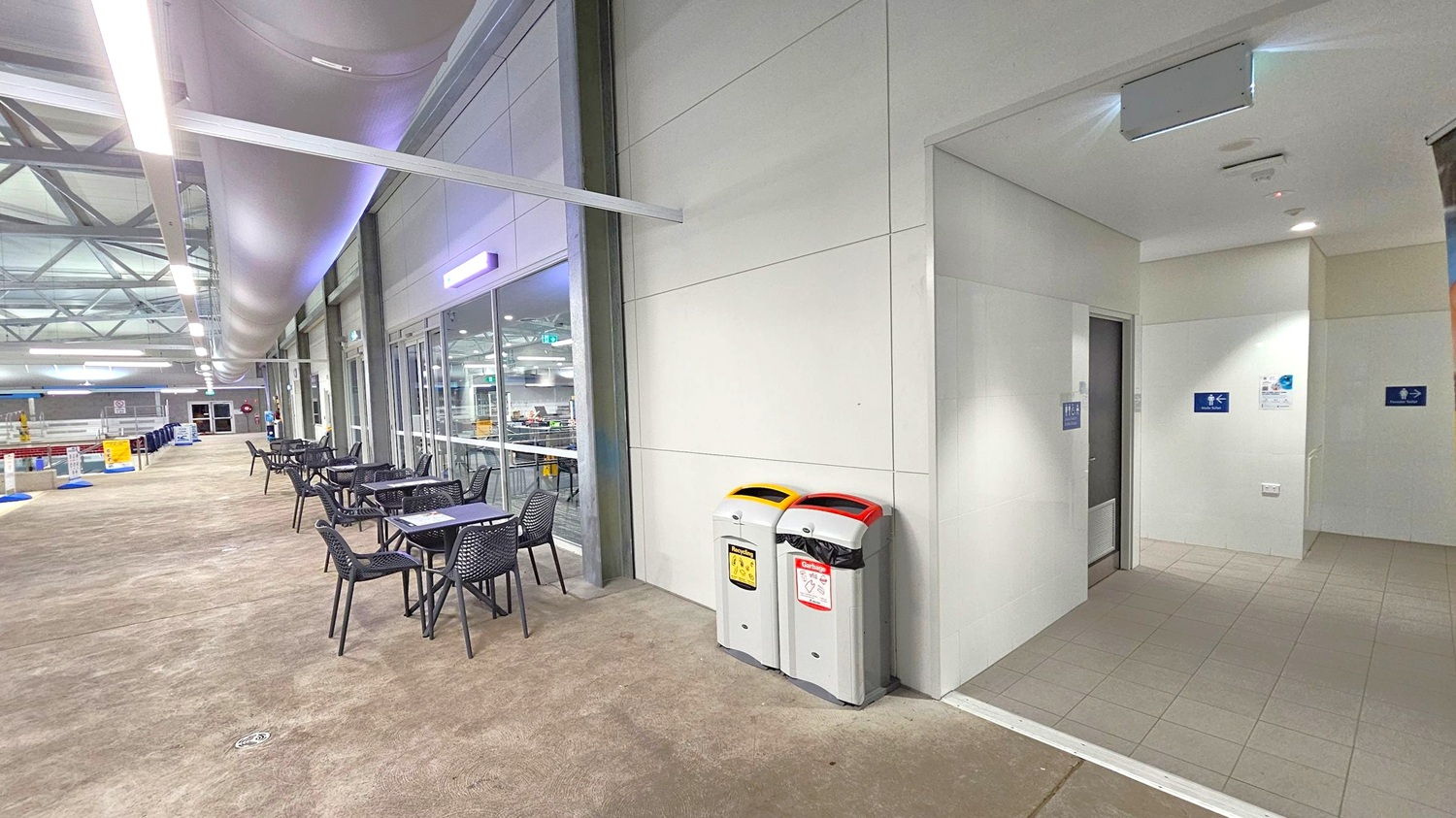 On the left, there are several black tables and chairs arranged along a glass wall, with a view into an indoor area. The floor is a light brown, textured surface. On the right, there are two bins with yellow and red lids next to a white tiled entrance leading to restrooms, indicated by signs on the wall. The ceiling features exposed ductwork and lighting, with a modern industrial design. On the left, there are several black tables and chairs arranged along a glass wall, with a view into an indoor area. The floor is a light brown, textured surface. On the right, there are two bins with yellow and red lids next to a white tiled entrance leading to restrooms, indicated by signs on the wall. The ceiling features exposed ductwork and lighting, with a modern industrial design.