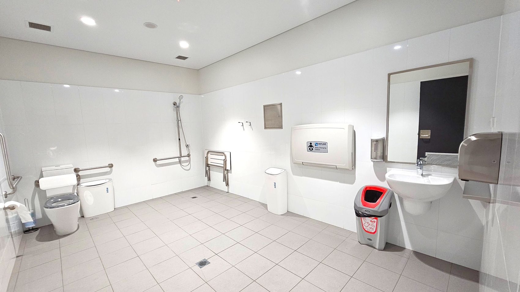 A spacious, accessible bathroom with white tiled walls and grey floor tiles. On the left, there is a toilet with support rails and a shower area with a handheld showerhead. In the centre, a foldable seat is mounted on the wall, with a nappy changing station above it. On the right, a sink with a mirror above it is accompanied by a soap dispenser and a hand dryer. A red and grey bin is positioned next to the sink. The ceiling is white with recessed lighting. A spacious, accessible bathroom with white tiled walls and grey floor tiles. On the left, there is a toilet with support rails and a shower area with a handheld showerhead. In the centre, a foldable seat is mounted on the wall, with a nappy changing station above it. On the right, a sink with a mirror above it is accompanied by a soap dispenser and a hand dryer. A red and grey bin is positioned next to the sink. The ceiling is white with recessed lighting.