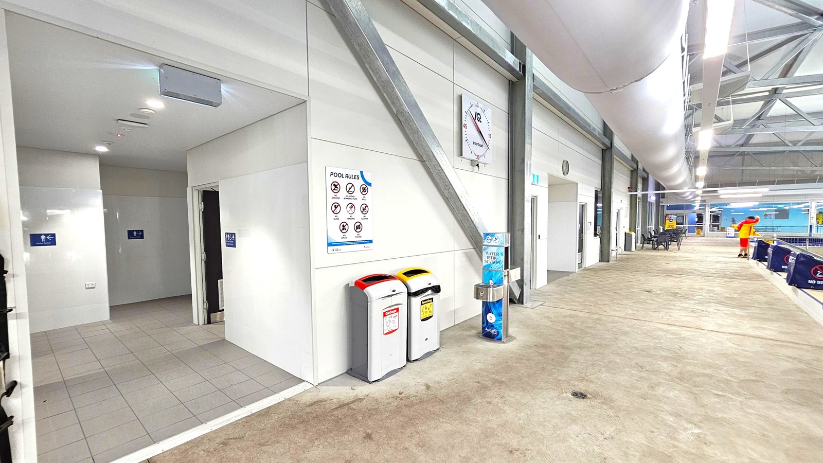 On the left, there are entrances to changing rooms with blue signs on white tiled walls. In the middle, a wall displays pool rules and a clock, with two bins below, one with a red lid and the other with a yellow lid. A water station is nearby. On the right, a corridor with tables and chairs leads to a pool area, where a person in a yellow and red outfit stands. The floor is a mix of green and brown, and the ceiling has exposed beams and large white ducts. On the left, there are entrances to changing rooms with blue signs on white tiled walls. In the middle, a wall displays pool rules and a clock, with two bins below, one with a red lid and the other with a yellow lid. A water station is nearby. On the right, a corridor with tables and chairs leads to a pool area, where a person in a yellow and red outfit stands. The floor is a mix of green and brown, and the ceiling has exposed beams and large white ducts.