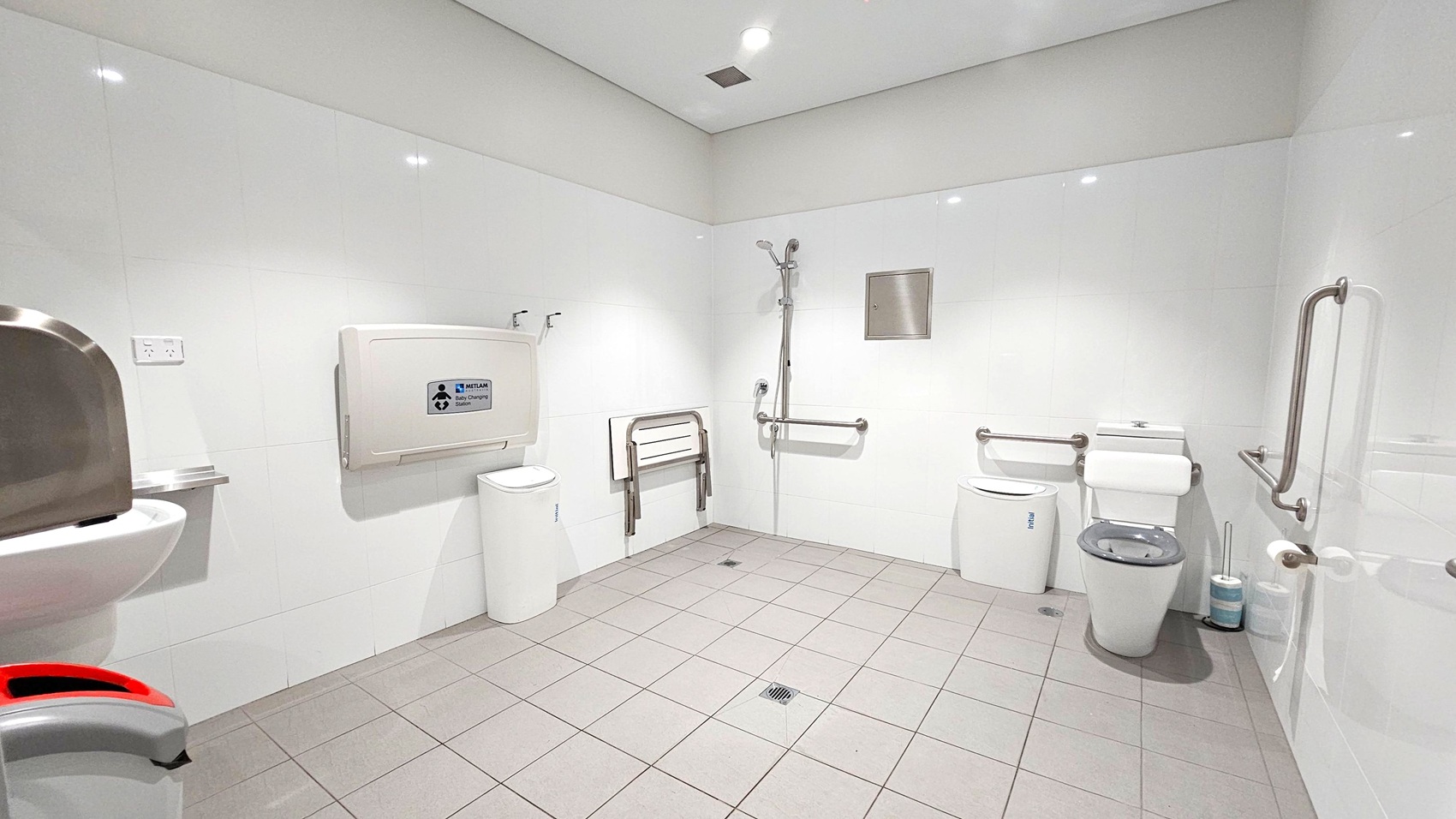 A spacious, accessible bathroom with white tiled walls and grey tiled floor. On the left, a sink with a soap dispenser and a hand dryer. In the centre, a baby changing station and a foldable seat attached to the wall. A shower with a handheld nozzle is also present. On the right, a toilet with support rails and a sanitary bin. The ceiling is white with recessed lighting. A spacious, accessible bathroom with white tiled walls and grey tiled floor. On the left, a sink with a soap dispenser and a hand dryer. In the centre, a baby changing station and a foldable seat attached to the wall. A shower with a handheld nozzle is also present. On the right, a toilet with support rails and a sanitary bin. The ceiling is white with recessed lighting.