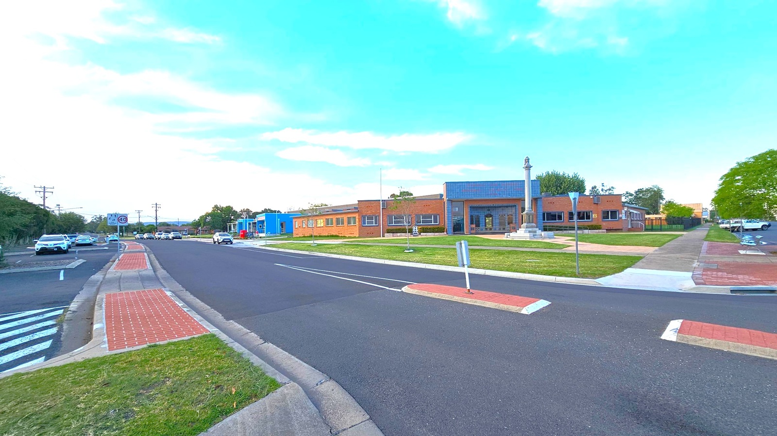 A wide street with a central median and a pedestrian crossing. On the left, several cars are parked along the road, with a speed limit sign indicating 40. On the right, a grassy area with a monument featuring a tall column. Behind it, a series of brick buildings with large windows. The pavement is a mix of red bricks and concrete, and there are trees in the background. The sky is clear with some clouds. A wide street with a central median and a pedestrian crossing. On the left, several cars are parked along the road, with a speed limit sign indicating 40. On the right, a grassy area with a monument featuring a tall column. Behind it, a series of brick buildings with large windows. The pavement is a mix of red bricks and concrete, and there are trees in the background. The sky is clear with some clouds.