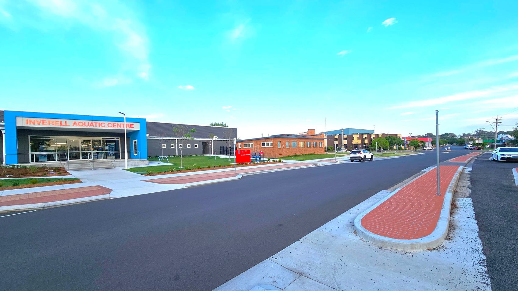 On the left, a building with a blue facade and large windows is labelled "Inverell Aquatic Centre". In the middle, a wide road with a red brick pedestrian island runs parallel to the building. On the right, a white car is parked along the kerb, and another vehicle is driving down the road. The background features a mix of modern and older buildings, with some greenery visible. The pavement is concrete, and the sky is clear. On the left, a building with a blue facade and large windows is labelled "Inverell Aquatic Centre". In the middle, a wide road with a red brick pedestrian island runs parallel to the building. On the right, a white car is parked along the kerb, and another vehicle is driving down the road. The background features a mix of modern and older buildings, with some greenery visible. The pavement is concrete, and the sky is clear.