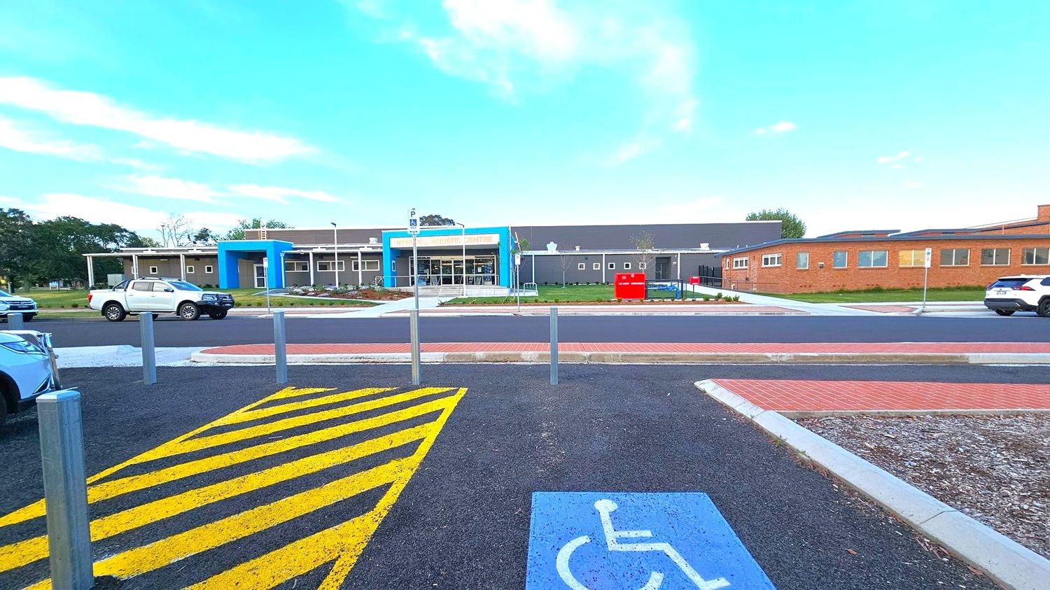 A building with a blue entrance and a sign above it is in the centre. The structure is surrounded by a paved area with a few parked cars, including a white pickup truck. In the foreground, there is a parking space marked with a blue disabled symbol and yellow diagonal lines. The ground is a mix of asphalt and brick paving. Trees and grass are visible in the background. A building with a blue entrance and a sign above it is in the centre. The structure is surrounded by a paved area with a few parked cars, including a white pickup truck. In the foreground, there is a parking space marked with a blue disabled symbol and yellow diagonal lines. The ground is a mix of asphalt and brick paving. Trees and grass are visible in the background.