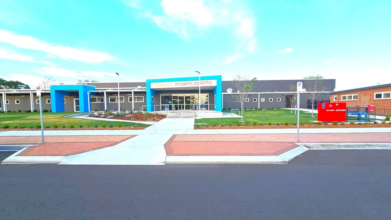 A modern building with a blue entrance and a sign reading "Inverell Aquatic Centre" in orange letters. The structure is surrounded by a well-maintained lawn and landscaped garden beds. A pathway leads from the street to the entrance. On the right, there is a red sign and some equipment. The foreground features a paved area and a road with a no-parking sign. The building has a flat roof and several small windows. A modern building with a blue entrance and a sign reading "Inverell Aquatic Centre" in orange letters. The structure is surrounded by a well-maintained lawn and landscaped garden beds. A pathway leads from the street to the entrance. On the right, there is a red sign and some equipment. The foreground features a paved area and a road with a no-parking sign. The building has a flat roof and several small windows.