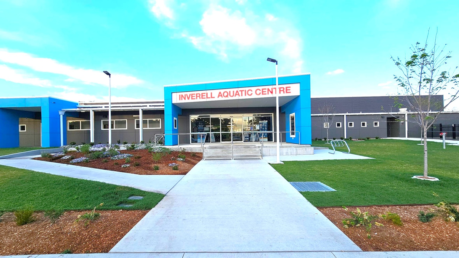 A modern building with a blue and grey facade, featuring a sign that reads "Inverell Aquatic Centre" in red letters above the entrance. The entrance has glass doors and is flanked by two white lamp posts. In the foreground, a concrete path leads to the entrance, bordered by landscaped gardens with small plants and mulch. On the right, there is a grassy area with young trees. The building has a flat roof and several small windows. A modern building with a blue and grey facade, featuring a sign that reads "Inverell Aquatic Centre" in red letters above the entrance. The entrance has glass doors and is flanked by two white lamp posts. In the foreground, a concrete path leads to the entrance, bordered by landscaped gardens with small plants and mulch. On the right, there is a grassy area with young trees. The building has a flat roof and several small windows.