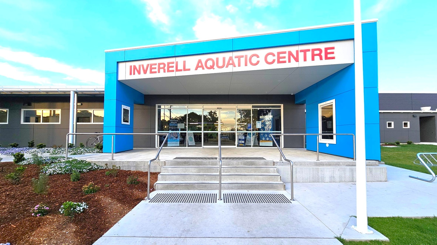 The entrance of the Inverell Aquatic Centre features a modern design with a blue and grey facade. Large glass doors are centrally located, flanked by windows. A set of concrete steps with metal railings leads up to the entrance. On the left, there is a landscaped area with small plants and flowers, and a bicycle is parked nearby. On the right, a bike rack is visible on the concrete path. The building has a flat roof, and the area is well-maintained. The entrance of the Inverell Aquatic Centre features a modern design with a blue and grey facade. Large glass doors are centrally located, flanked by windows. A set of concrete steps with metal railings leads up to the entrance. On the left, there is a landscaped area with small plants and flowers, and a bicycle is parked nearby. On the right, a bike rack is visible on the concrete path. The building has a flat roof, and the area is well-maintained.