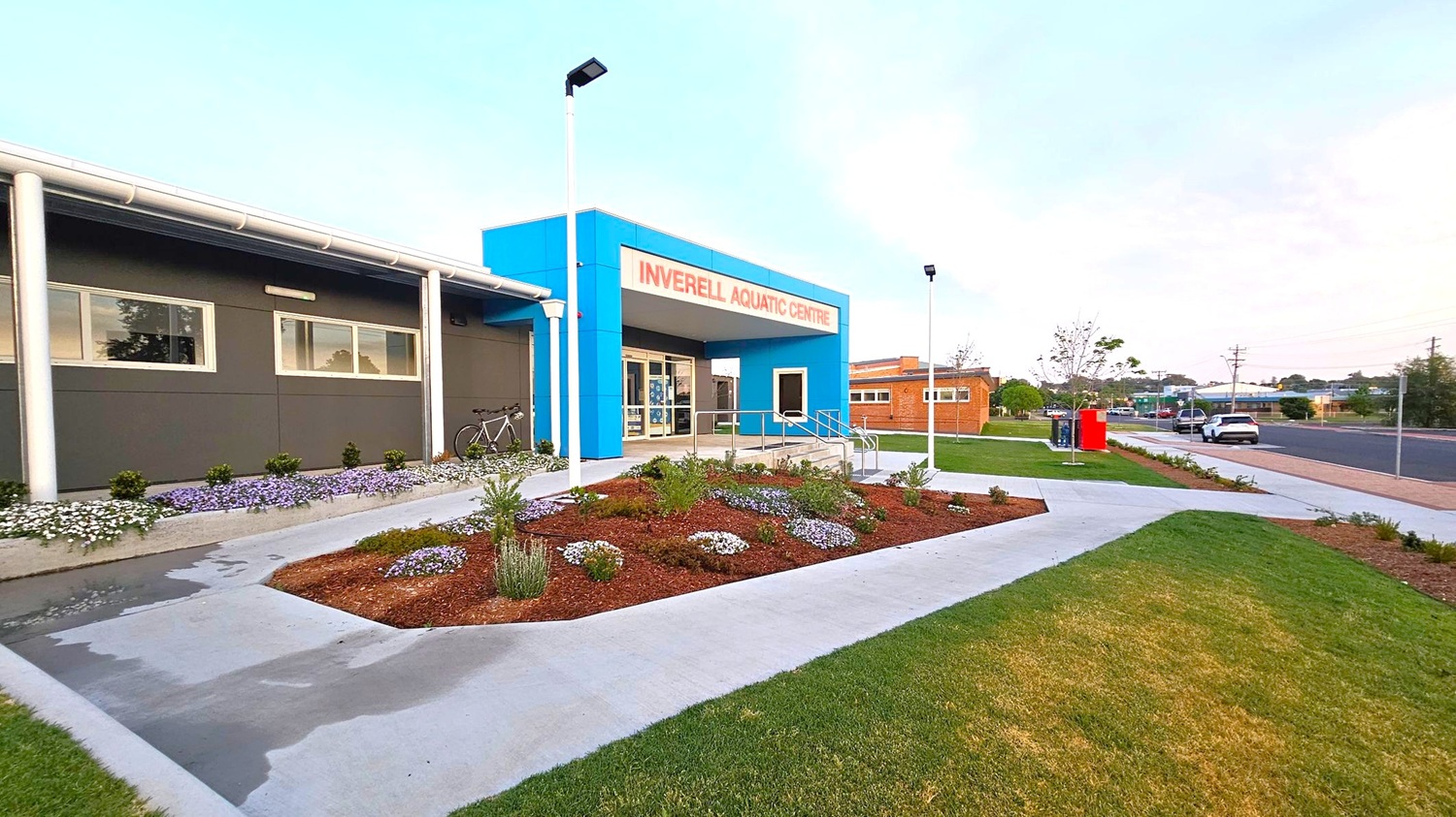 A modern building with a blue entrance labelled "Inverell Aquatic Centre" in red. The left side features a grey wall with windows and a bicycle parked nearby. In front, there is a landscaped garden with various plants and flowers. The right side shows a pathway leading to a street with a parked car and a red utility box. The area is well-lit with street lamps, and the ground is a mix of concrete paths and grassy patches. A modern building with a blue entrance labelled "Inverell Aquatic Centre" in red. The left side features a grey wall with windows and a bicycle parked nearby. In front, there is a landscaped garden with various plants and flowers. The right side shows a pathway leading to a street with a parked car and a red utility box. The area is well-lit with street lamps, and the ground is a mix of concrete paths and grassy patches.