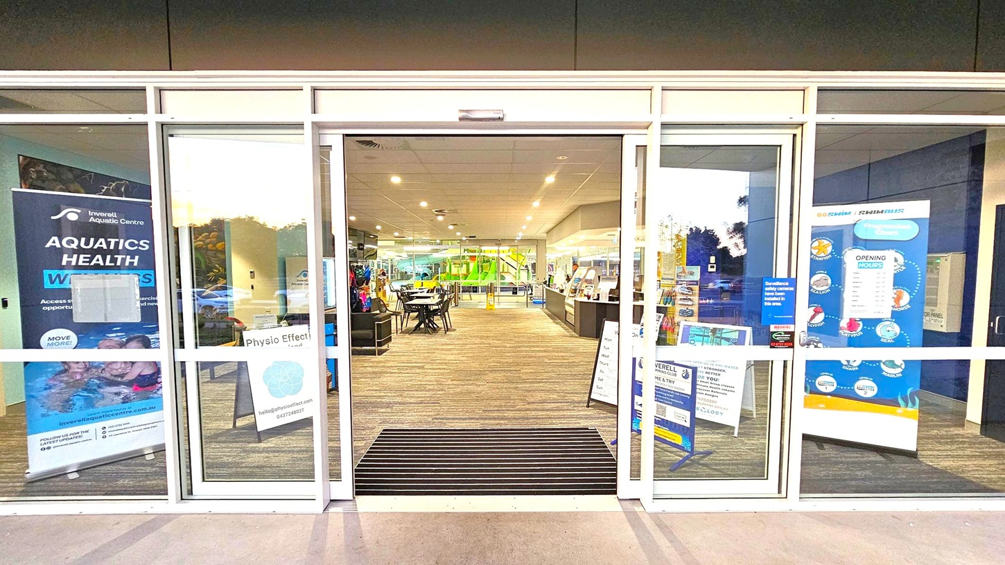 Glass entrance doors to a facility with signs on both sides. On the left, a banner for an aquatic centre and a sign for "Physio Effect." On the right, a poster with opening hours and a progression chart. Inside, a carpeted floor leads to tables and chairs, with a green slide visible in the background. The ceiling is fitted with recessed lighting. Glass entrance doors to a facility with signs on both sides. On the left, a banner for an aquatic centre and a sign for "Physio Effect." On the right, a poster with opening hours and a progression chart. Inside, a carpeted floor leads to tables and chairs, with a green slide visible in the background. The ceiling is fitted with recessed lighting.