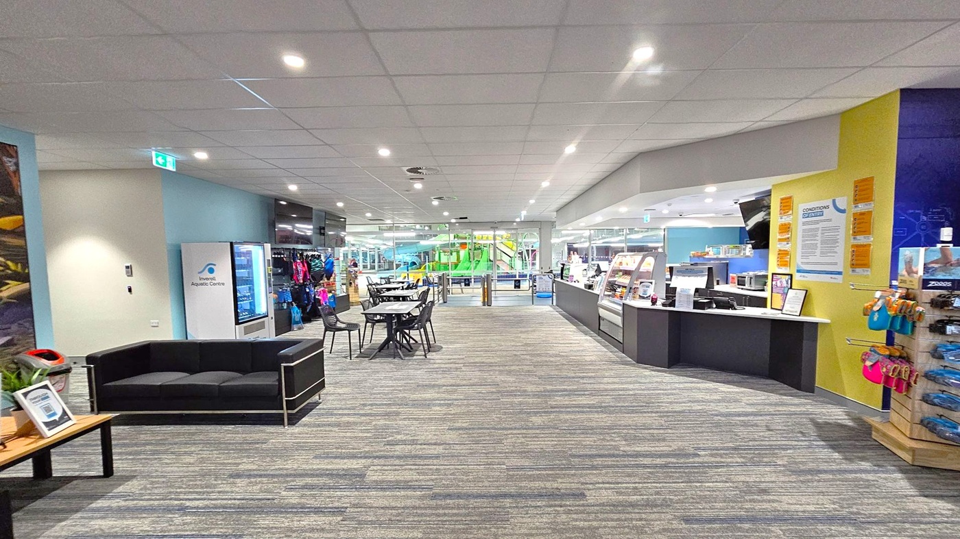 A spacious indoor area with a grey and green carpeted floor and a white panelled ceiling with recessed lighting. On the left, there's a black sofa and a vending machine labelled "Inverell Aquatic Centre." In the centre, tables and chairs are arranged, leading to glass doors revealing a green and blue water slide. On the right, a counter with a display case and a wall with notices and merchandise, including swim gear, is visible. A spacious indoor area with a grey and green carpeted floor and a white panelled ceiling with recessed lighting. On the left, there's a black sofa and a vending machine labelled "Inverell Aquatic Centre." In the centre, tables and chairs are arranged, leading to glass doors revealing a green and blue water slide. On the right, a counter with a display case and a wall with notices and merchandise, including swim gear, is visible.