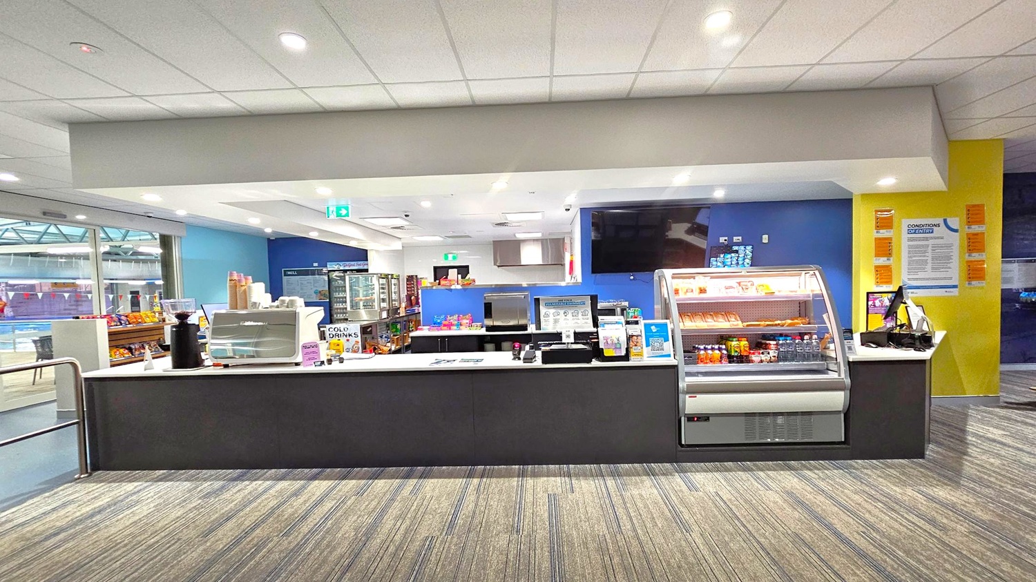 A snack bar with a counter displaying various items. On the left, there are shelves with snacks and a coffee machine. In the centre, a refrigerated display case contains drinks and snacks. Behind the counter, a menu board and a refrigerator with more items are visible. The right side has a yellow wall with informational posters. The floor is carpeted with a striped pattern, and the ceiling is white with recessed lighting. A snack bar with a counter displaying various items. On the left, there are shelves with snacks and a coffee machine. In the centre, a refrigerated display case contains drinks and snacks. Behind the counter, a menu board and a refrigerator with more items are visible. The right side has a yellow wall with informational posters. The floor is carpeted with a striped pattern, and the ceiling is white with recessed lighting.