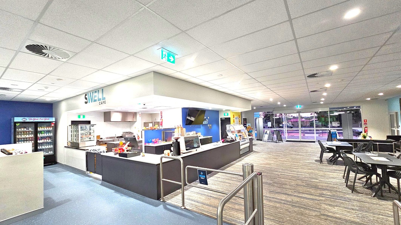 A café interior with a counter in the centre displaying snacks and a coffee machine. The sign above reads "Swell Café". To the left, a refrigerated display holds drinks. On the right, there are tables and chairs on a carpeted floor. The ceiling is fitted with recessed lighting and an exit sign. Large windows in the background show an outdoor view. A café interior with a counter in the centre displaying snacks and a coffee machine. The sign above reads "Swell Café". To the left, a refrigerated display holds drinks. On the right, there are tables and chairs on a carpeted floor. The ceiling is fitted with recessed lighting and an exit sign. Large windows in the background show an outdoor view.